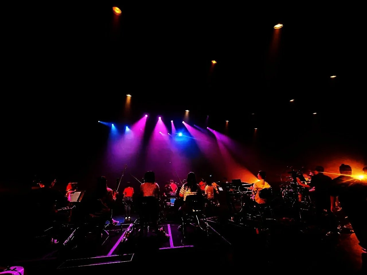 Silhouettes of musicians playing instruments on a dark stage illuminated by colorful purple, pink, blue, and orange lights.