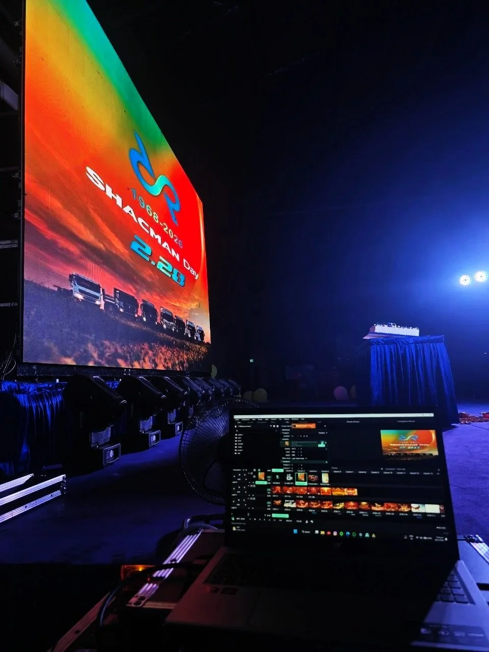 A large screen displays a colorful graphic with the text '1983-2023 Shachman Day 2.20'. In front, a laptop running video editing or broadcasting software is visible, with stage lights and equipment nearby in a dark setting.