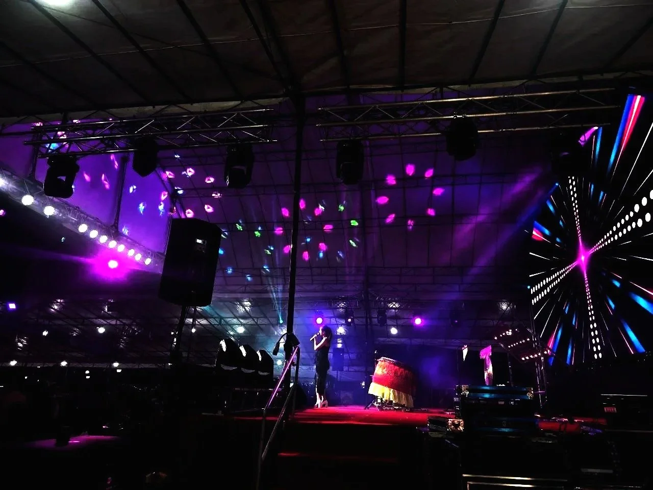 A performer singing on a stage with colorful lights and a large digital screen displaying a vibrant abstract pattern, under a high ceiling with dark metal framework.