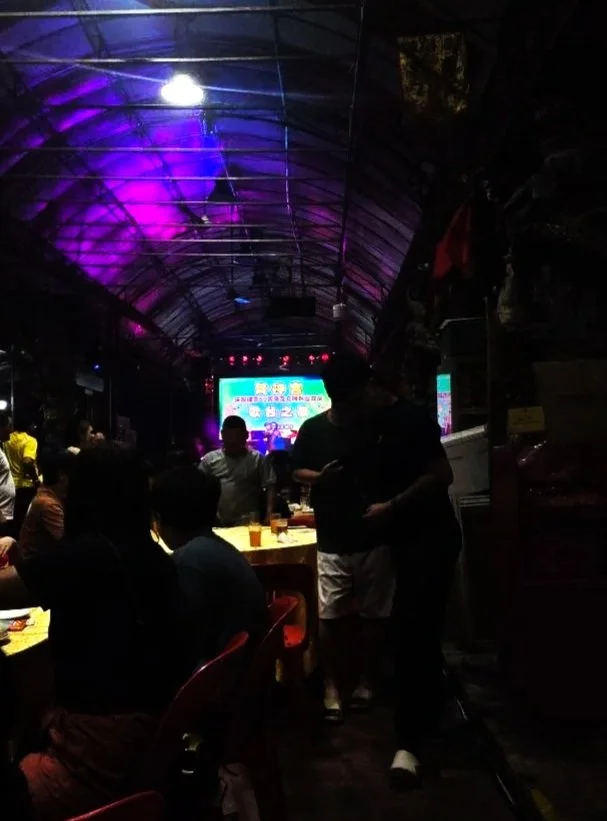 People sitting around tables in a dimly lit restaurant or bar with a stage and brightly lit screen in the background.