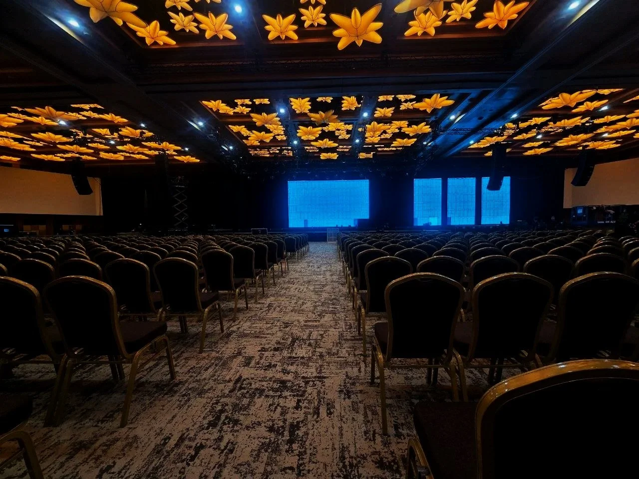 Empty conference or event hall with rows of chairs facing a stage with large screens, ceiling decorated with illuminated yellow flowers.