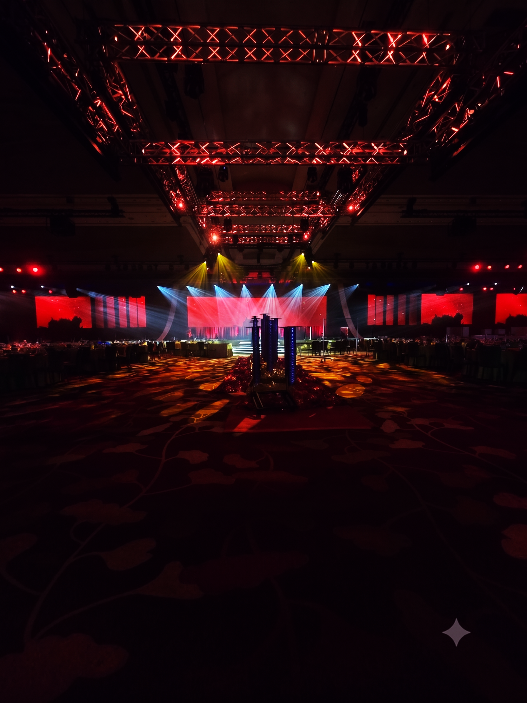 Colorful indoor event stage with bright red, blue, and yellow spotlights, large screens displaying red backgrounds, and decorative lighting effects on the floor.