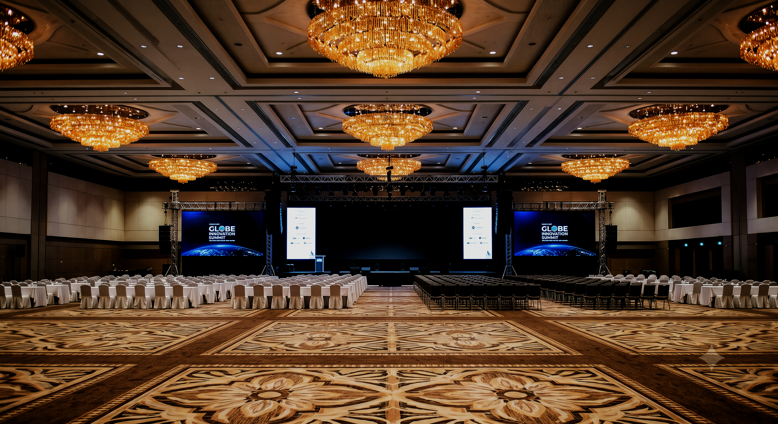 Empty conference hall with a stage, large screens displaying 'GLOBE INNOVATION SUMMIT,' rows of chairs and tables, and elegant chandeliers hanging from the ceiling.