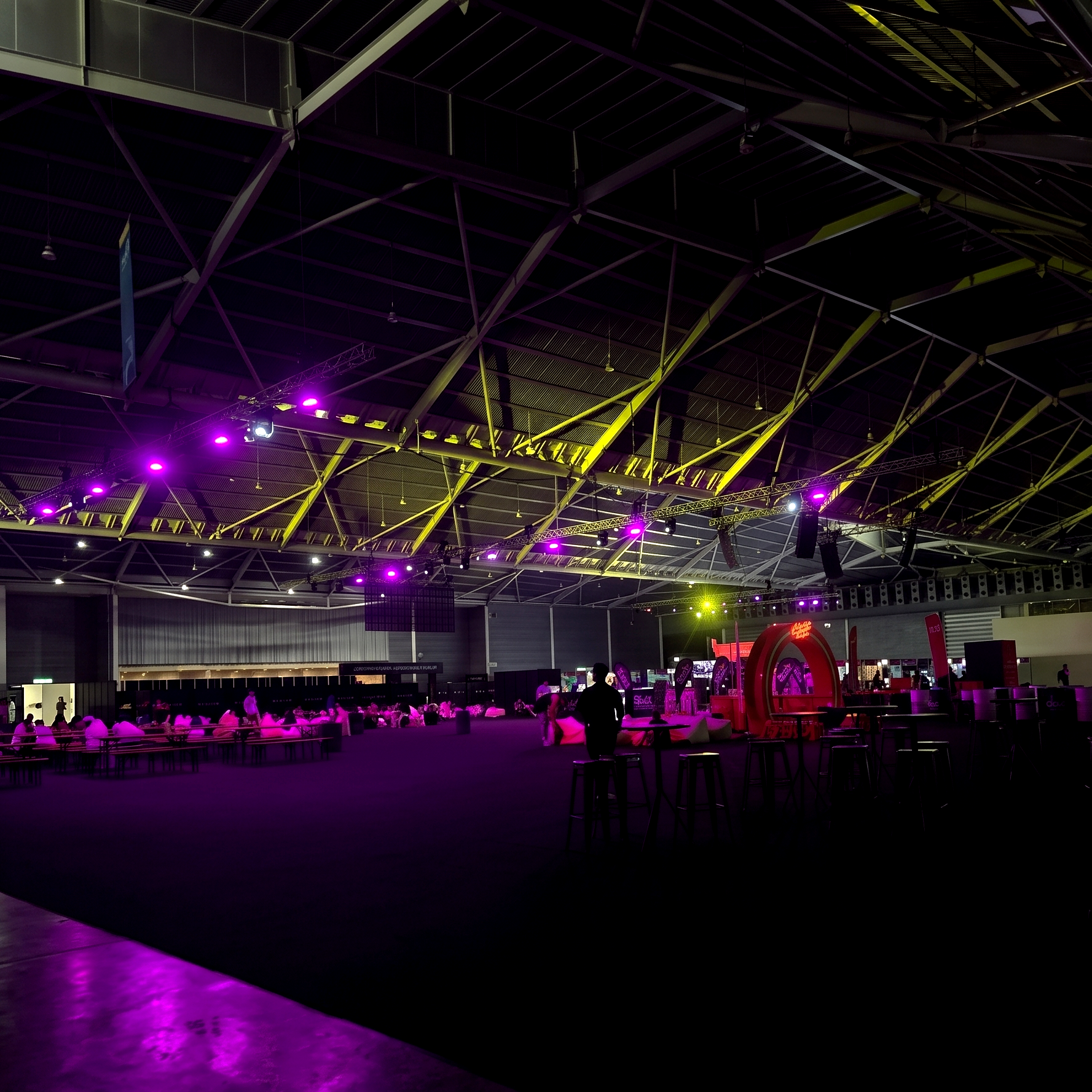 Interior of a large indoor event space with a high arched ceiling, illuminated with purple and yellow stage lights, people sitting and standing, some at tables, in a modern venue.