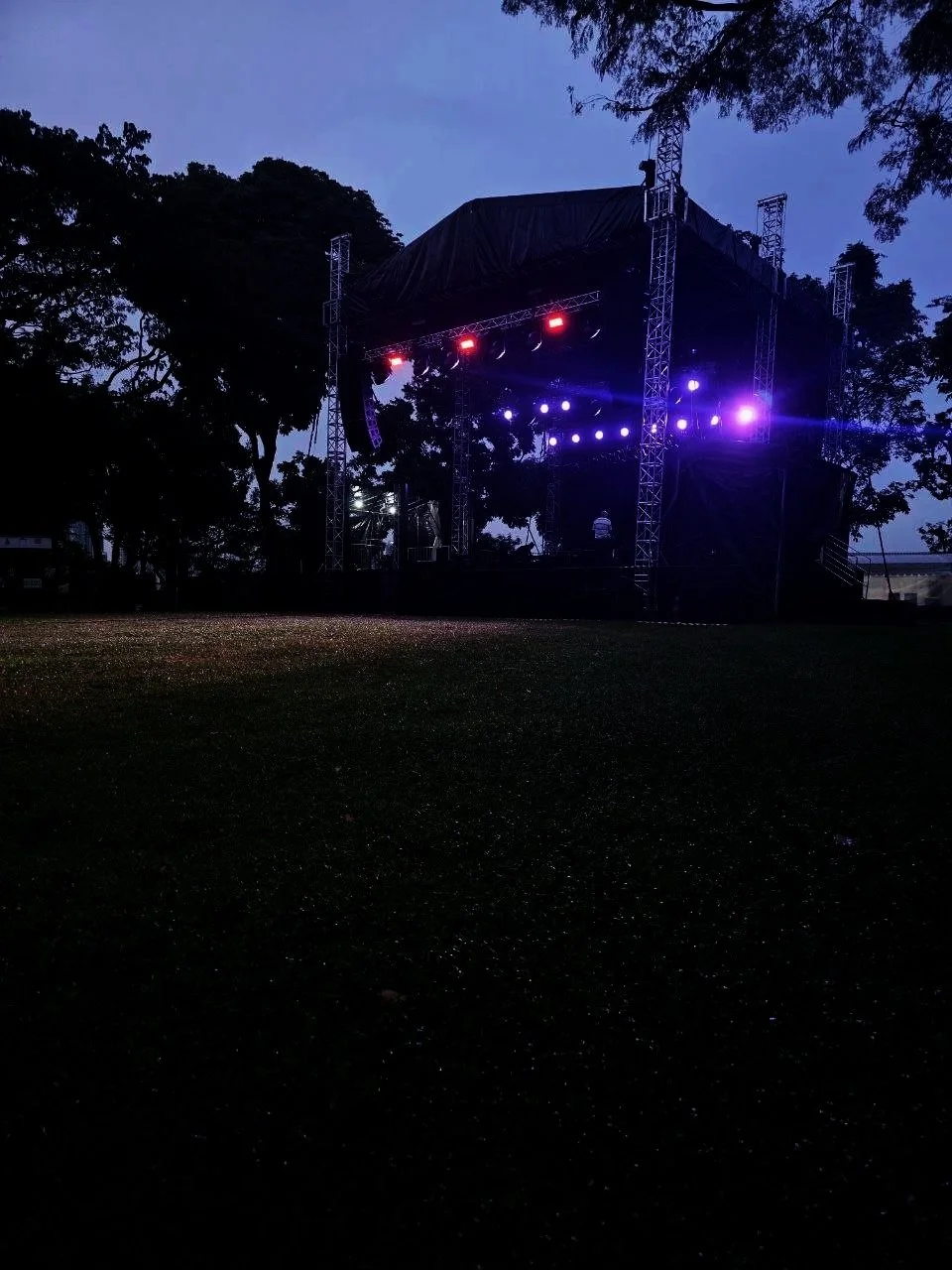 An outdoor music stage with lighting set up, surrounded by trees during dusk or early evening.