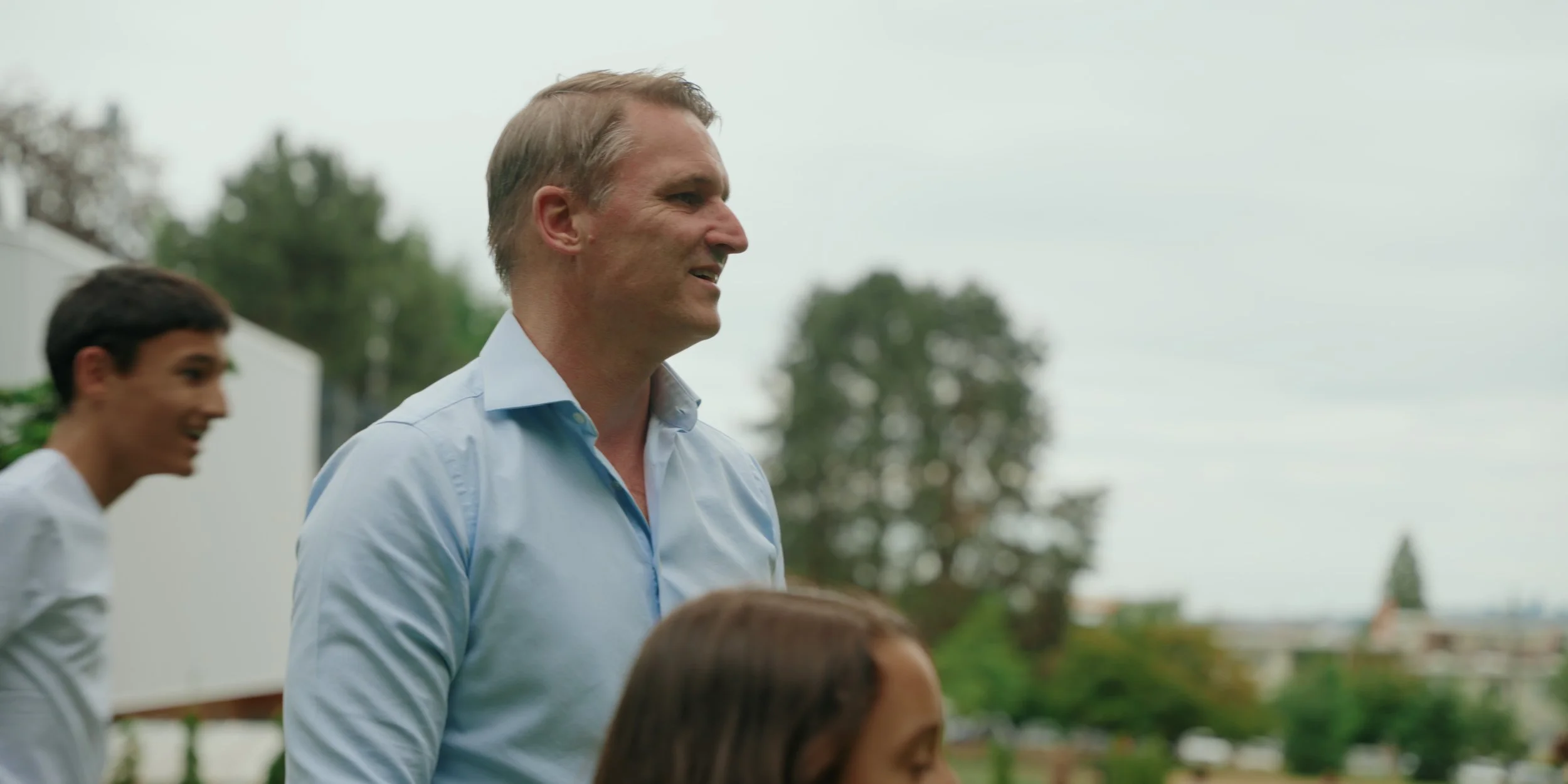 A group of people outdoors, with a man in a light blue button-up shirt in focus, smiling and looking to the right. Two other people are partially visible, a young man and a woman, both smiling.