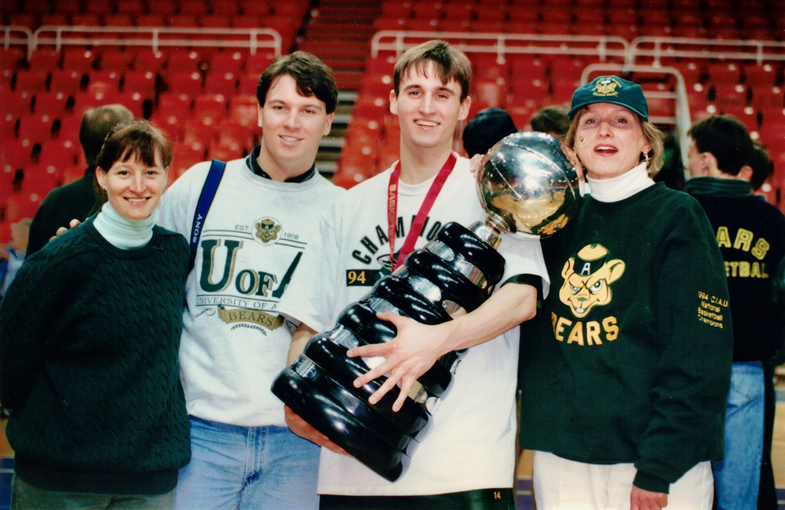 Group of four people, two young men and two women, celebrating with a trophy in an indoor sports arena with red seats in the background. The two women are wearing University of Florida Gators apparel.