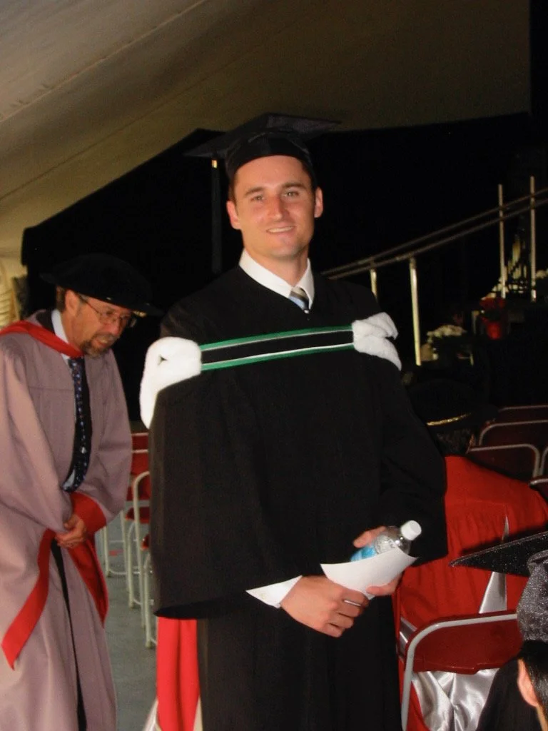 A young man in a graduation cap and gown holding a water bottle and a diploma, standing in an indoor auditorium with red chairs, next to an older man dressed in academic regalia.
