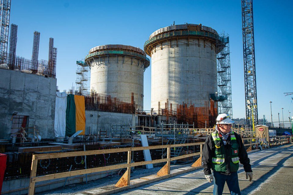 Construction worker wearing a helmet and safety vest at a building site with large concrete structures and a tower crane in the background under a clear blue sky.
