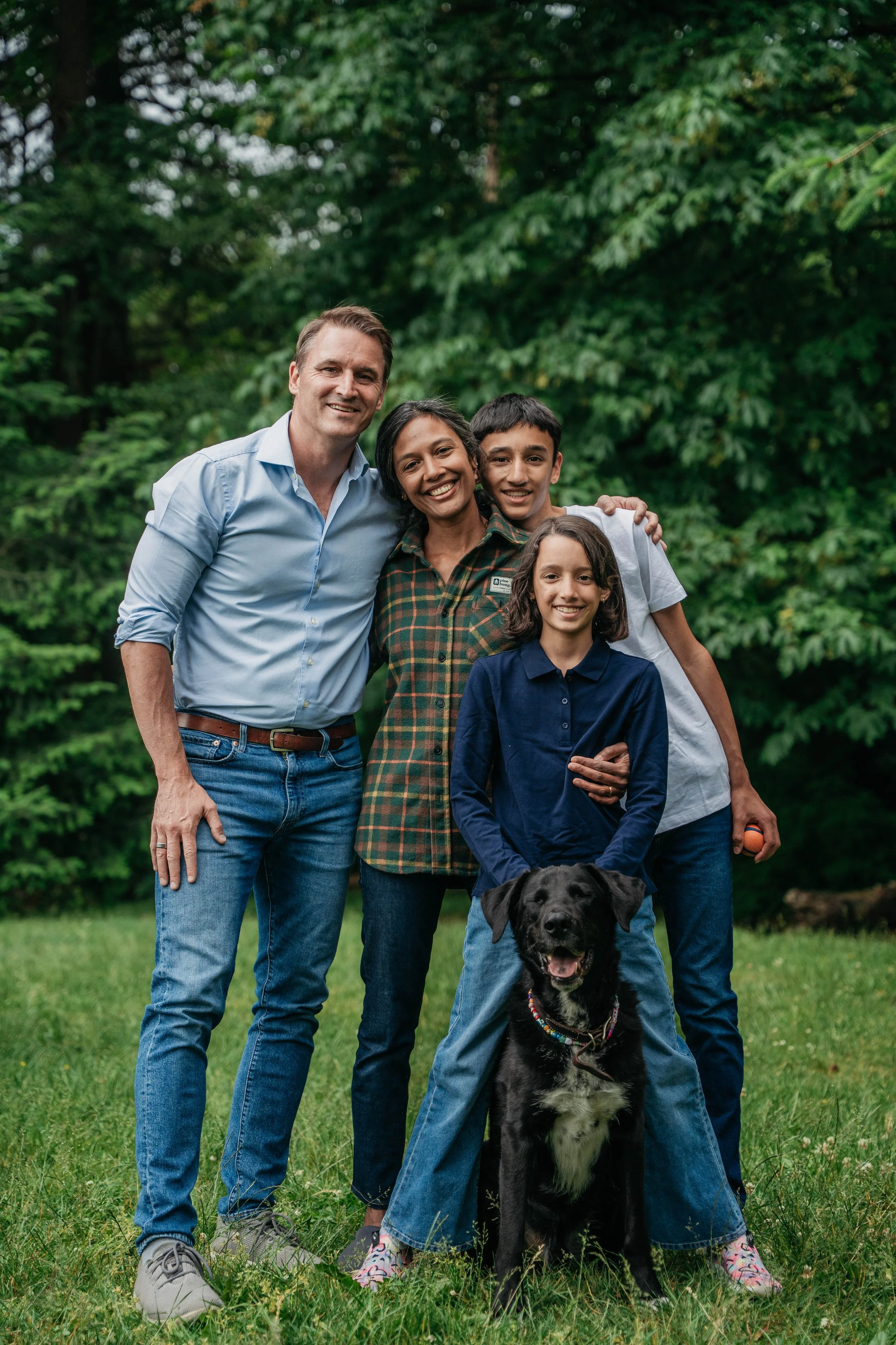 A group of four people, three children and one adult, standing outdoors on grass with trees in the background. They are smiling and close together, with a dog sitting in front of them.