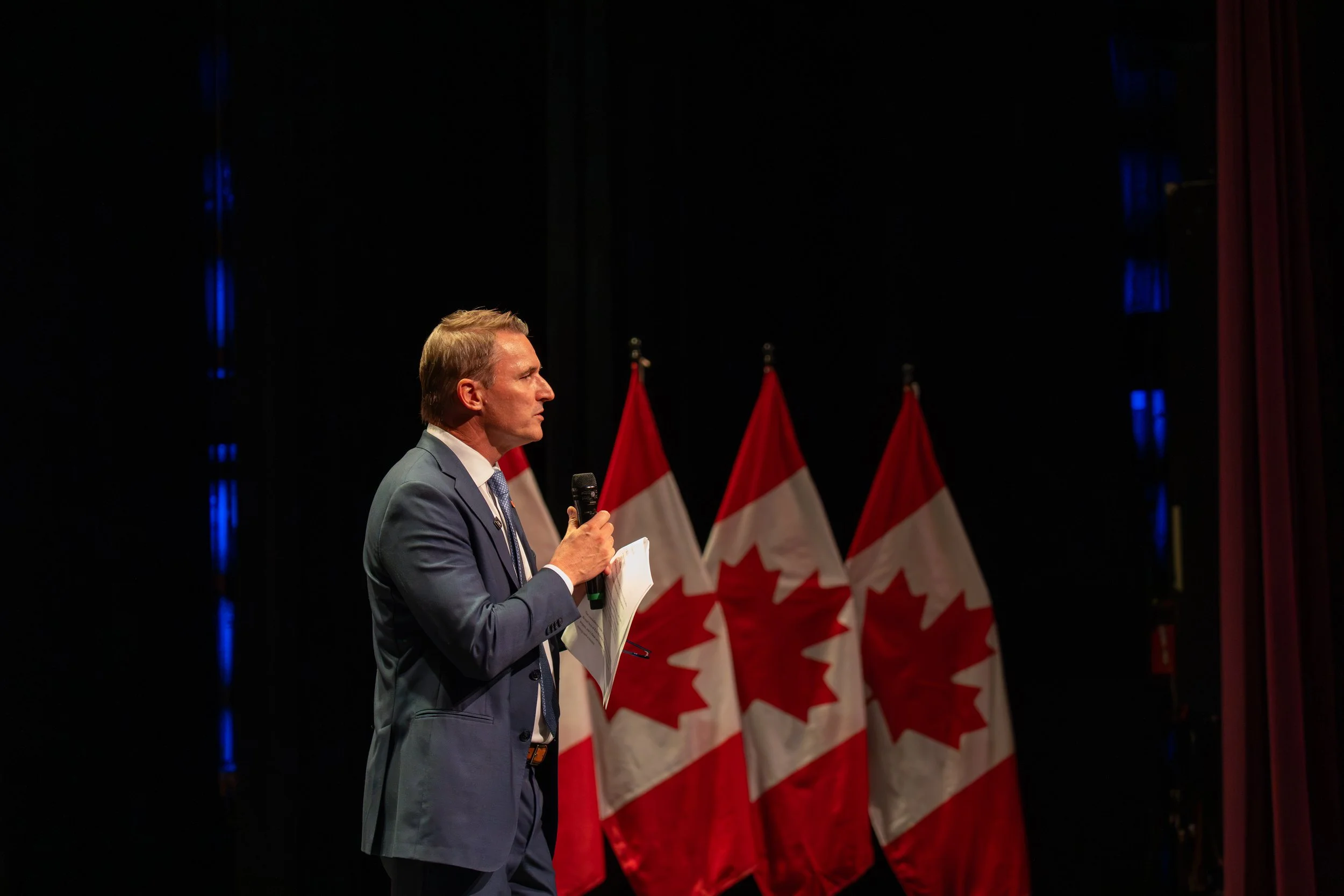 A man in a blue suit holding a microphone and reading from paper, standing in front of multiple Canadian flags on a stage.