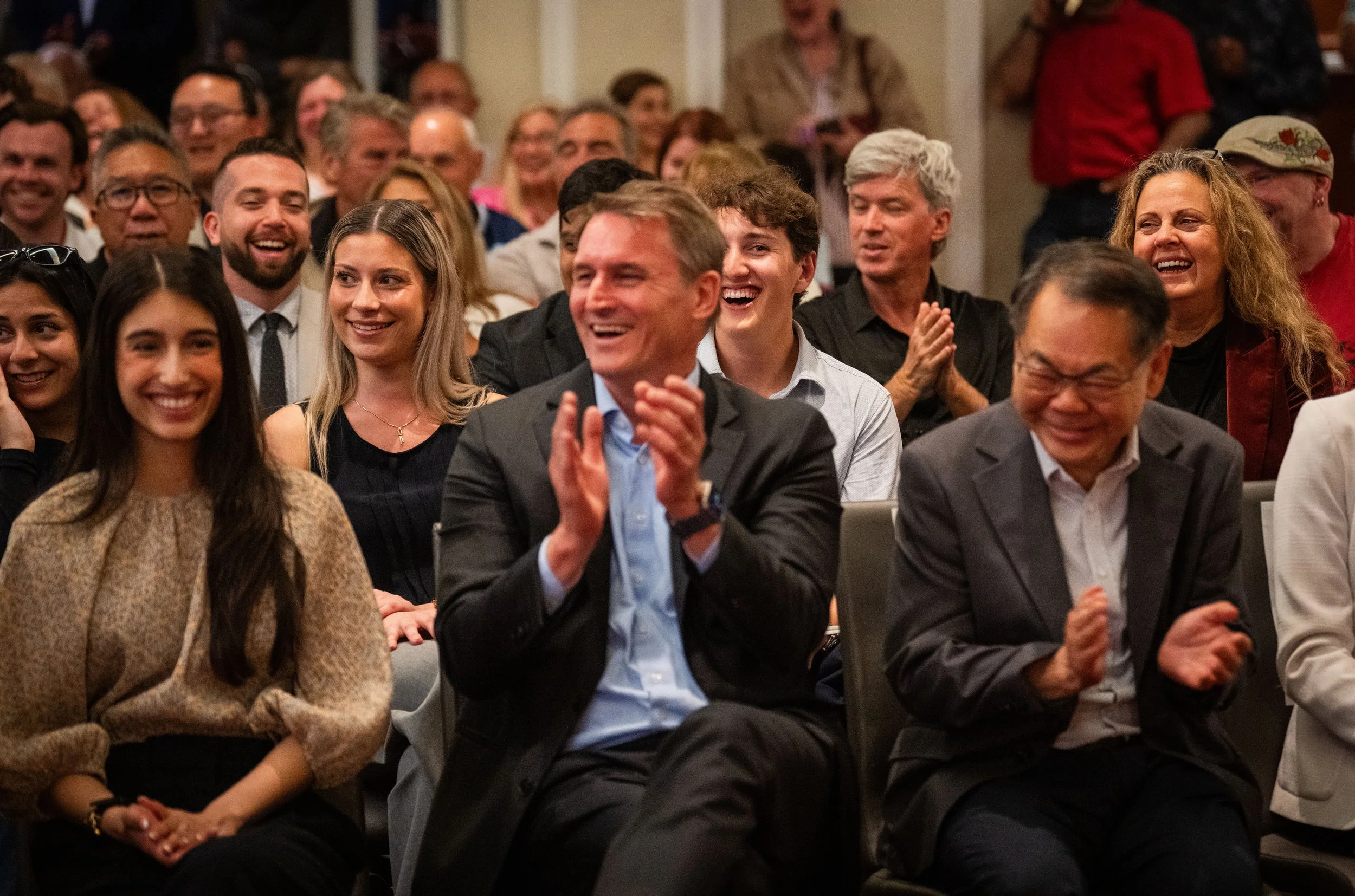 A group of people sitting in an audience, smiling and laughing at an event, with some clapping and showing expressions of joy.