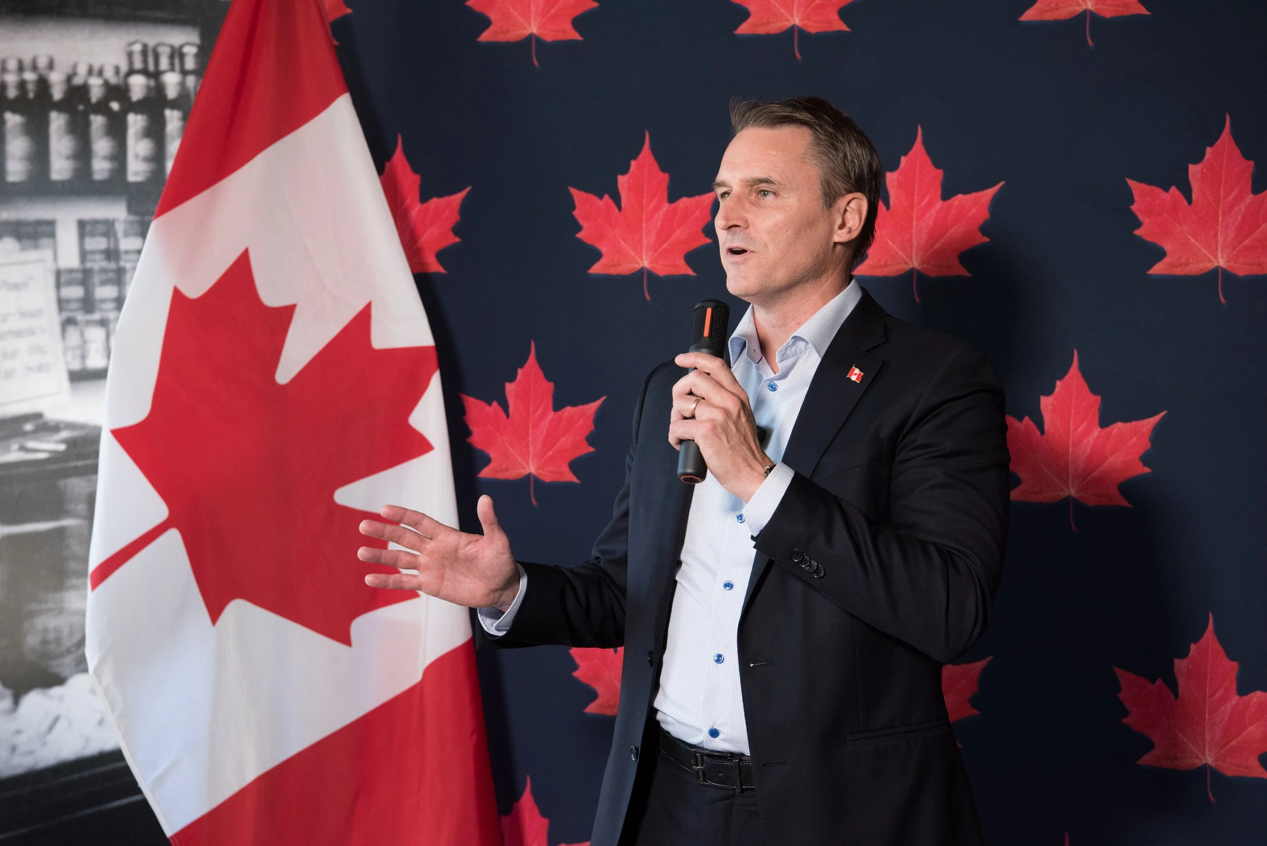 A man in a dark suit and light blue shirt speaking into a microphone, standing in front of a Canadian flag and a backdrop decorated with red maple leaves, suggesting a Canada-themed event or speech.