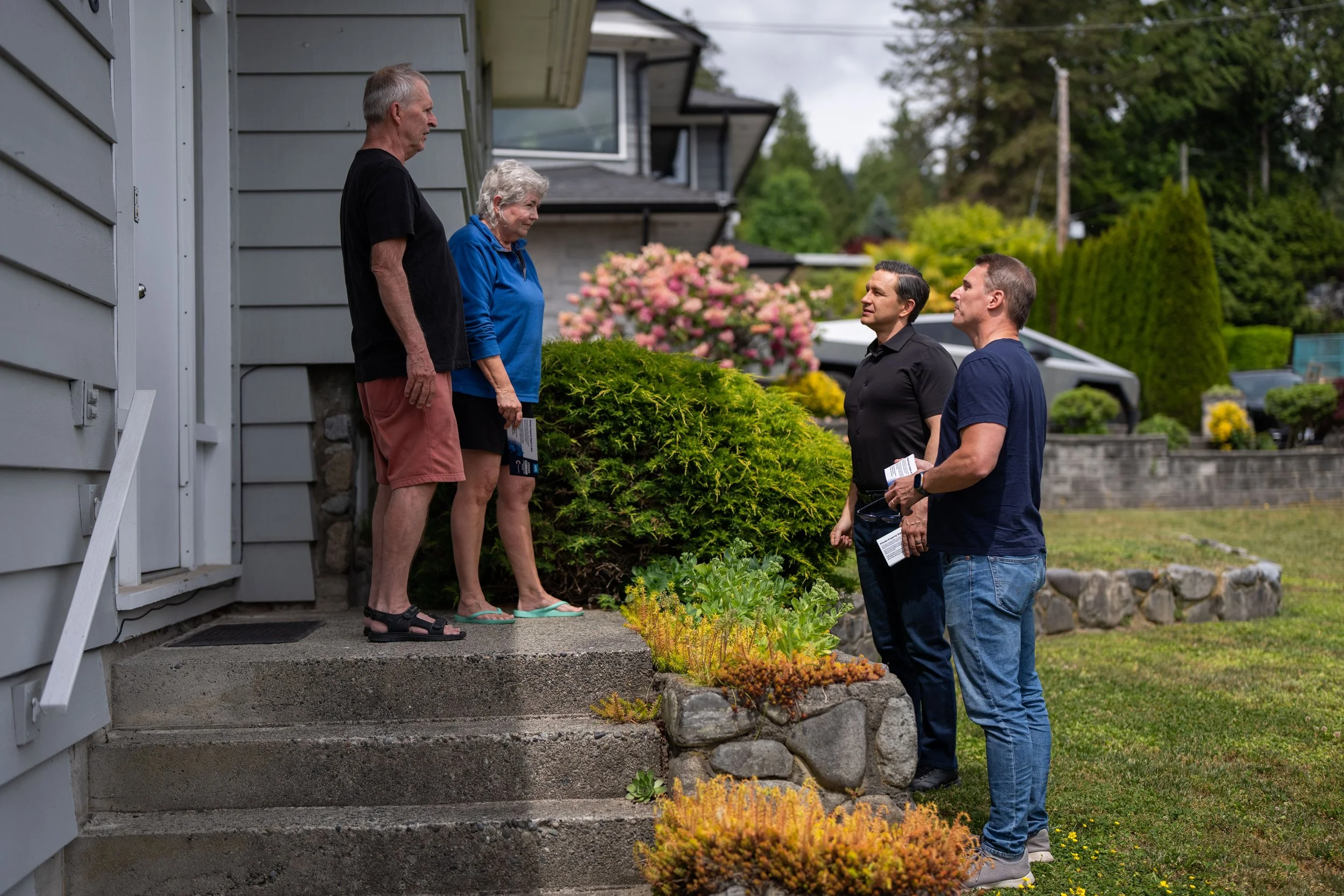 Five people having a conversation outside of a house, with two standing on the steps and three on the ground, surrounded by greenery and flowers.