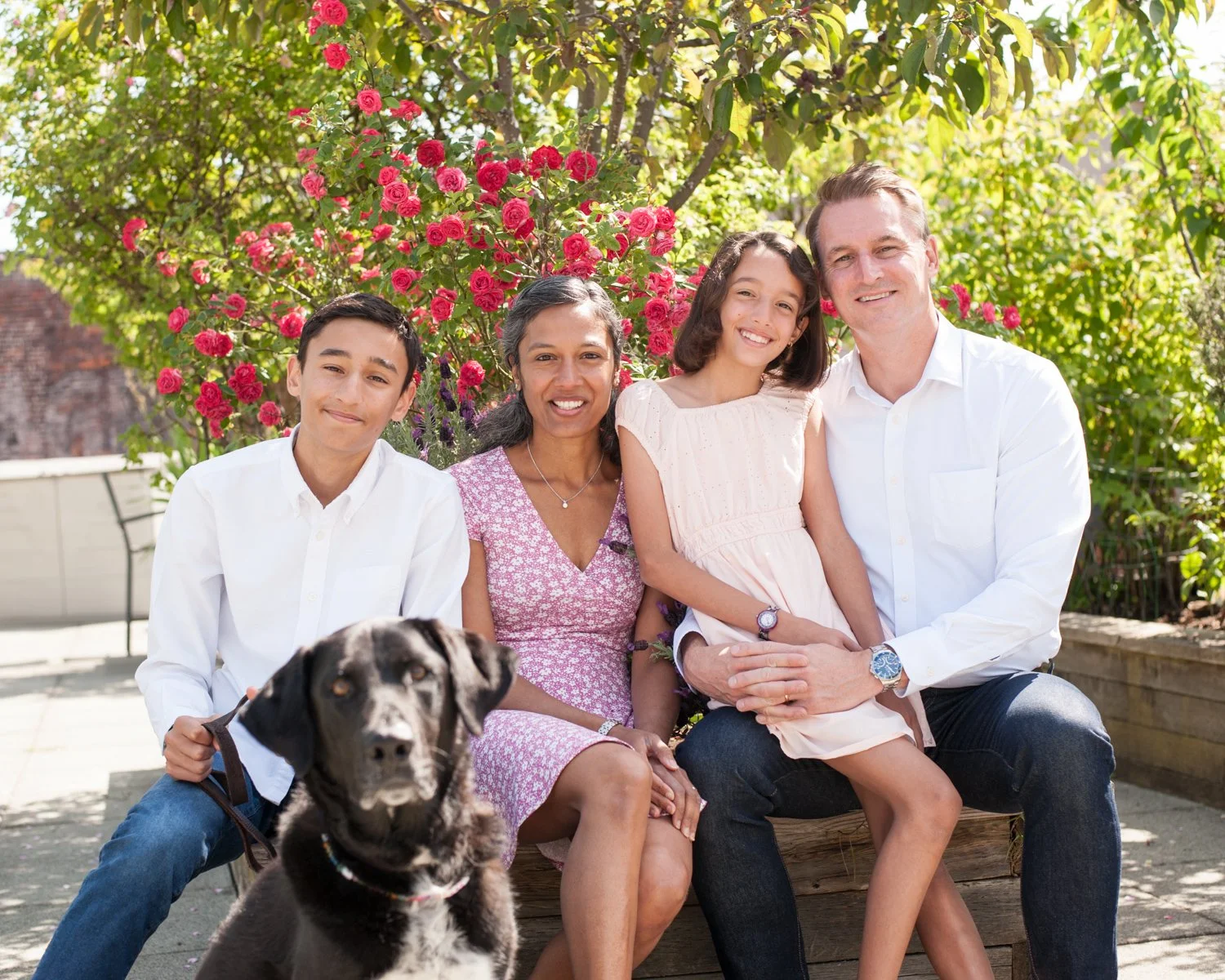A family of four sitting outdoors on a sunny day, with a dog in the foreground and pink flowering bushes in the background.