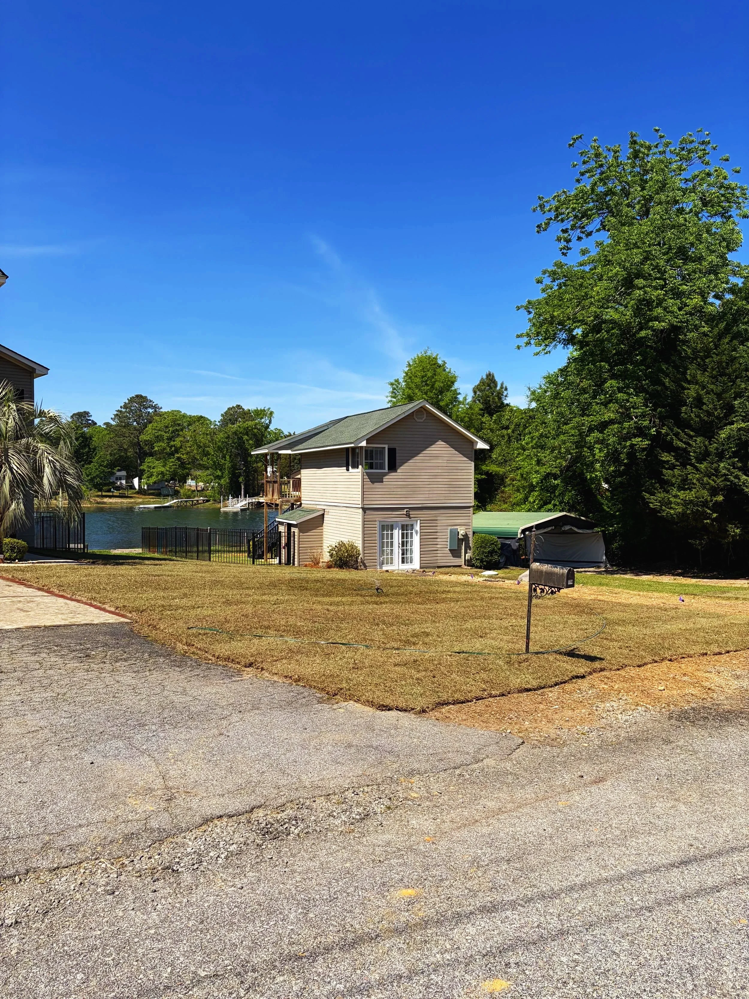 A residential area with a house by a body of water, surrounded by trees and greenery under a clear blue sky, with a paved driveway in the foreground.