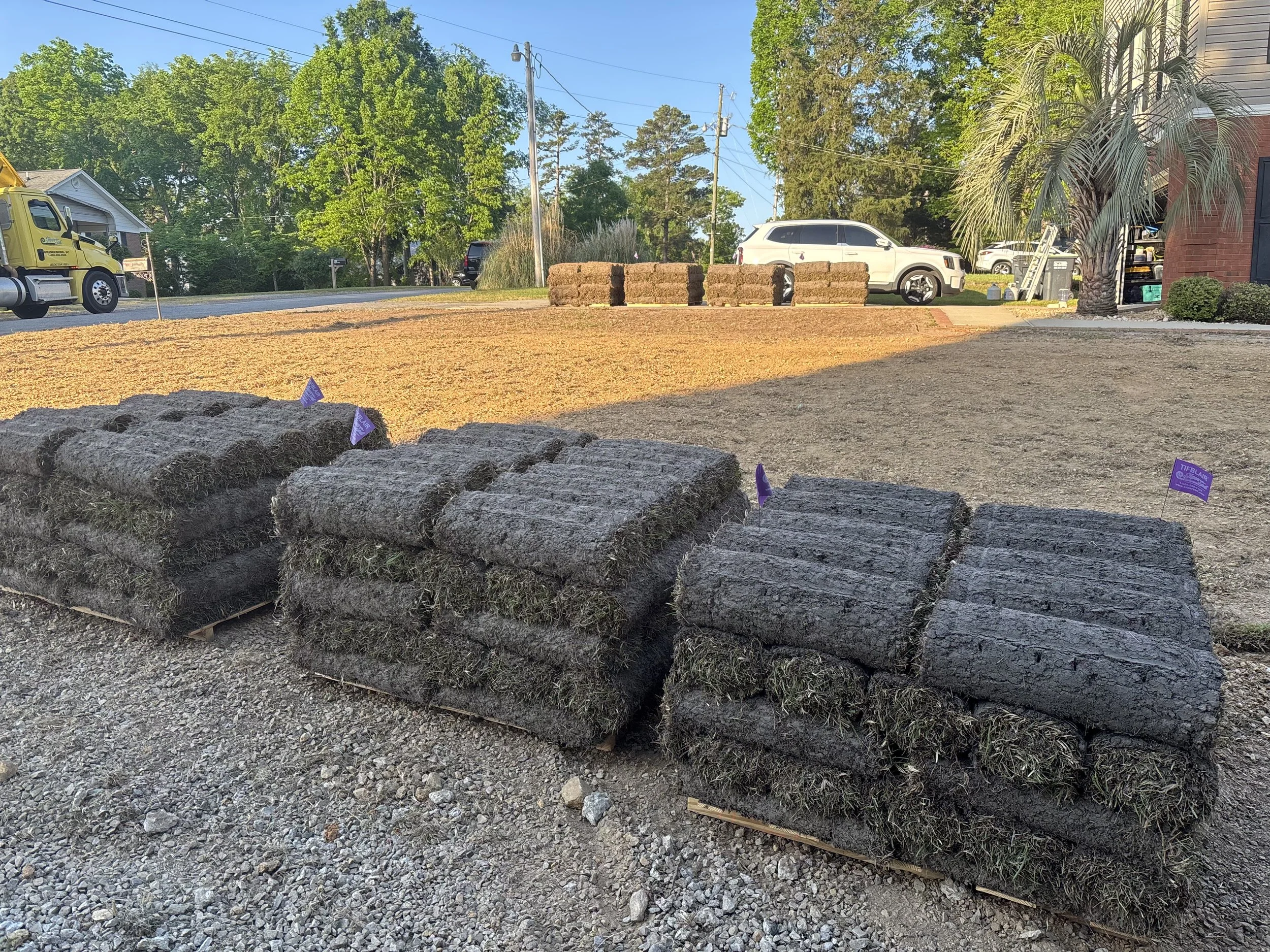 Construction site with soil and rocks spread on ground, stacks of bricks, construction flags, and parked vehicles including a white SUV and a yellow truck, with trees and a street in the background.