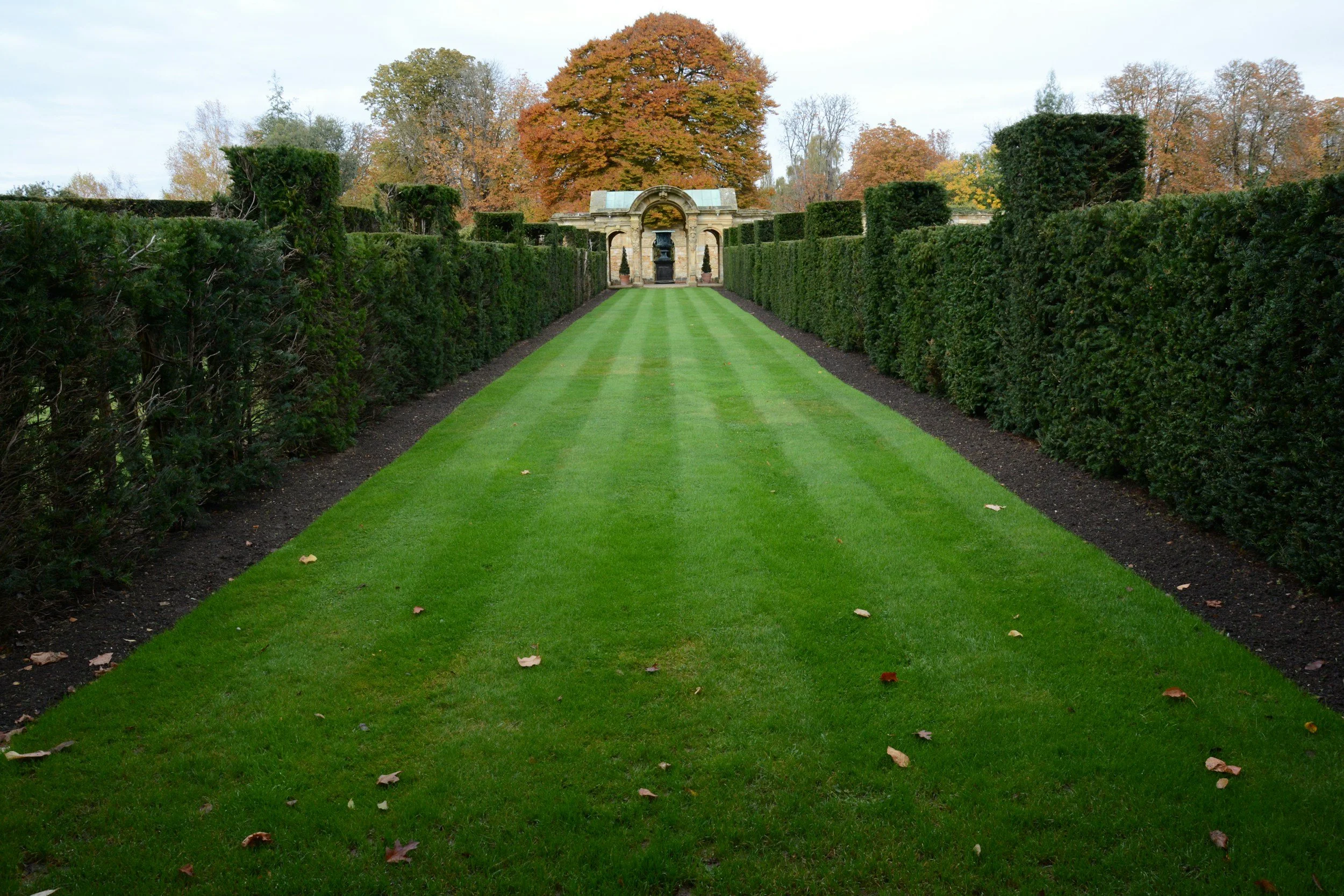A symmetrical garden with neatly trimmed hedges on both sides, a lush green lawn, and an ornate archway with a fountain in the background, surrounded by large trees with autumn-colored leaves.