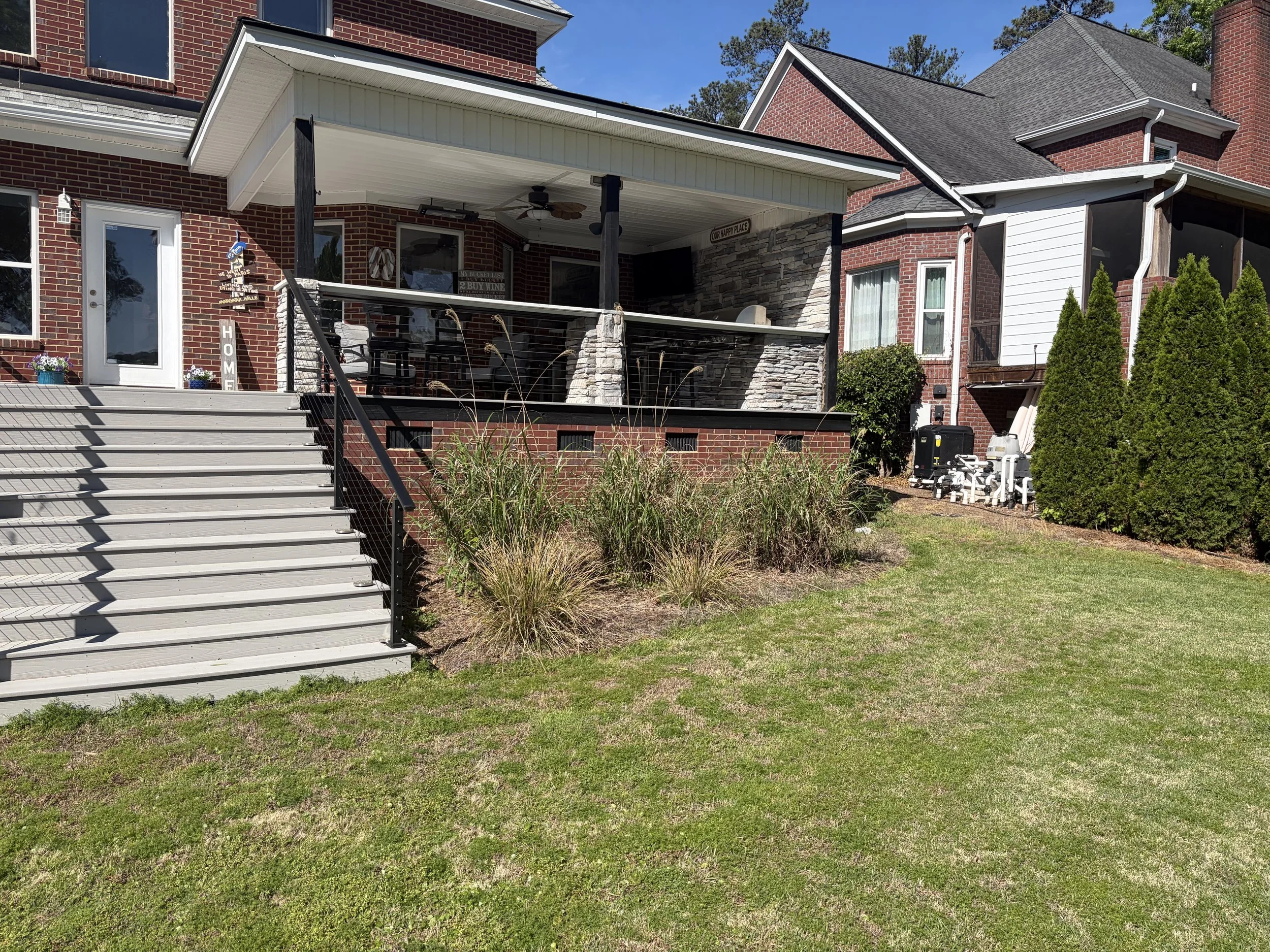 Backyard with stairs leading to a covered porch area with outdoor ceiling fans and decorative signs. Brick house with white siding and black trim, surrounded by green bushes and grass.