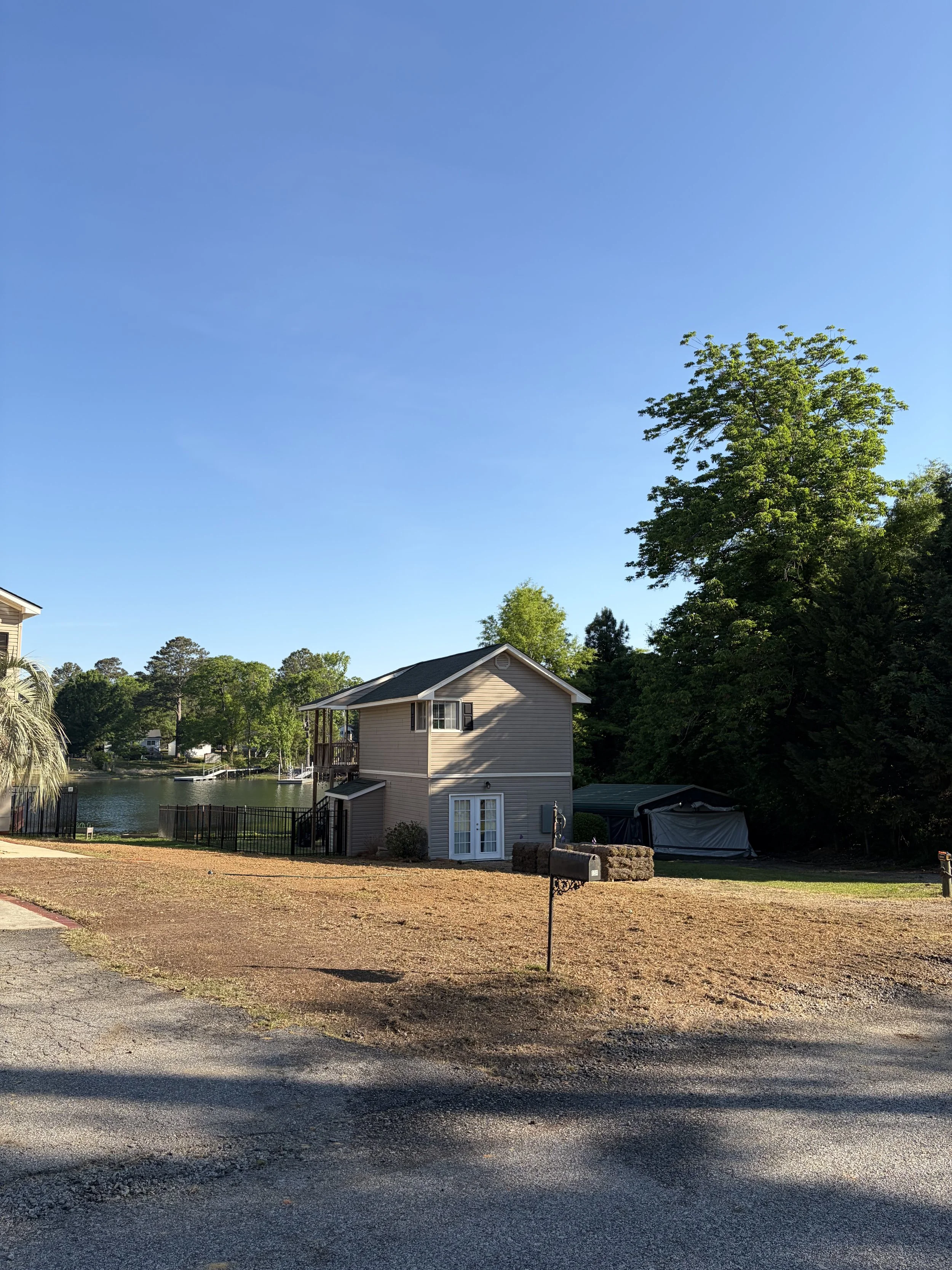 A two-story house near a lake with a backyard fence, surrounded by greenery and trees under a clear blue sky.