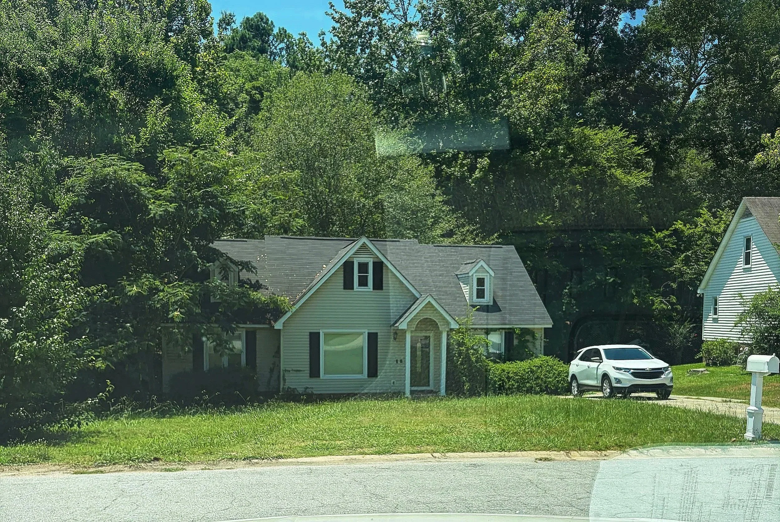 A suburban neighborhood view through a car window showing houses, parked cars, trees, and a street.