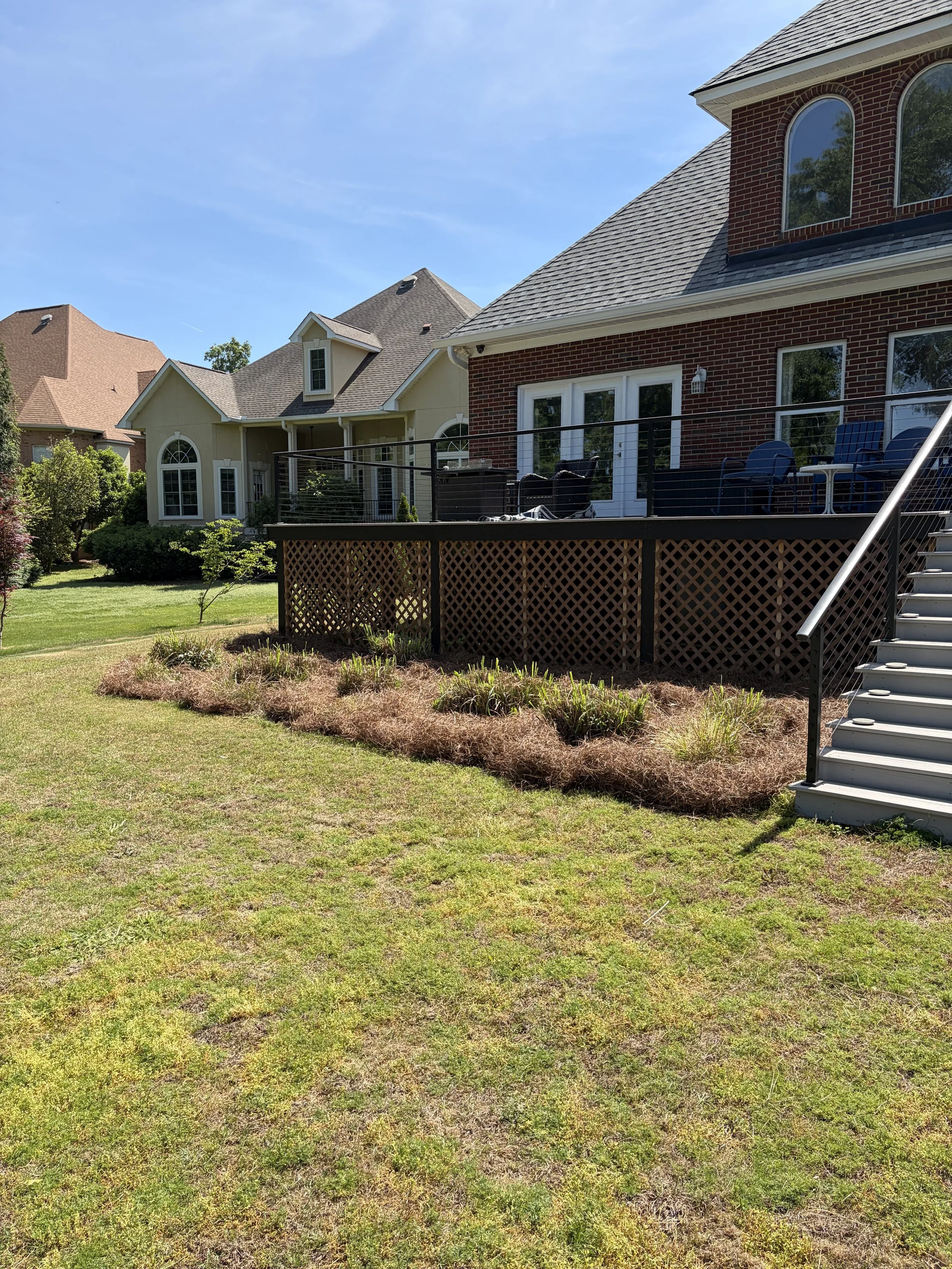 A backyard with a grassy lawn, a wooden deck with outdoor furniture, and a two-story brick house with large windows under a clear blue sky.