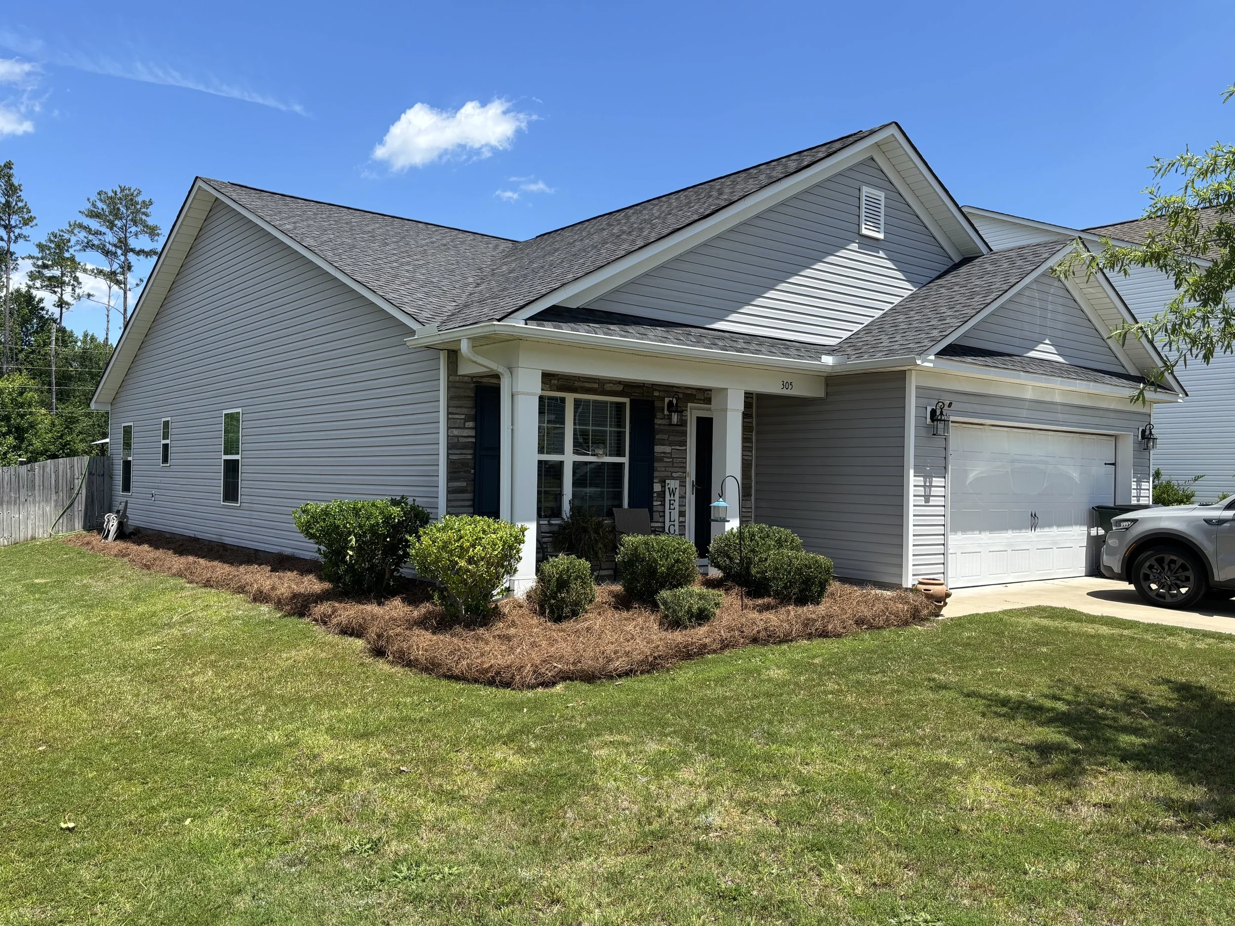 A modern suburban house with light gray siding, a dark gray shingled roof, and a white garage door. The front yard has a green lawn, bushes, and a small tree, with a blue sky and a few clouds in the background.