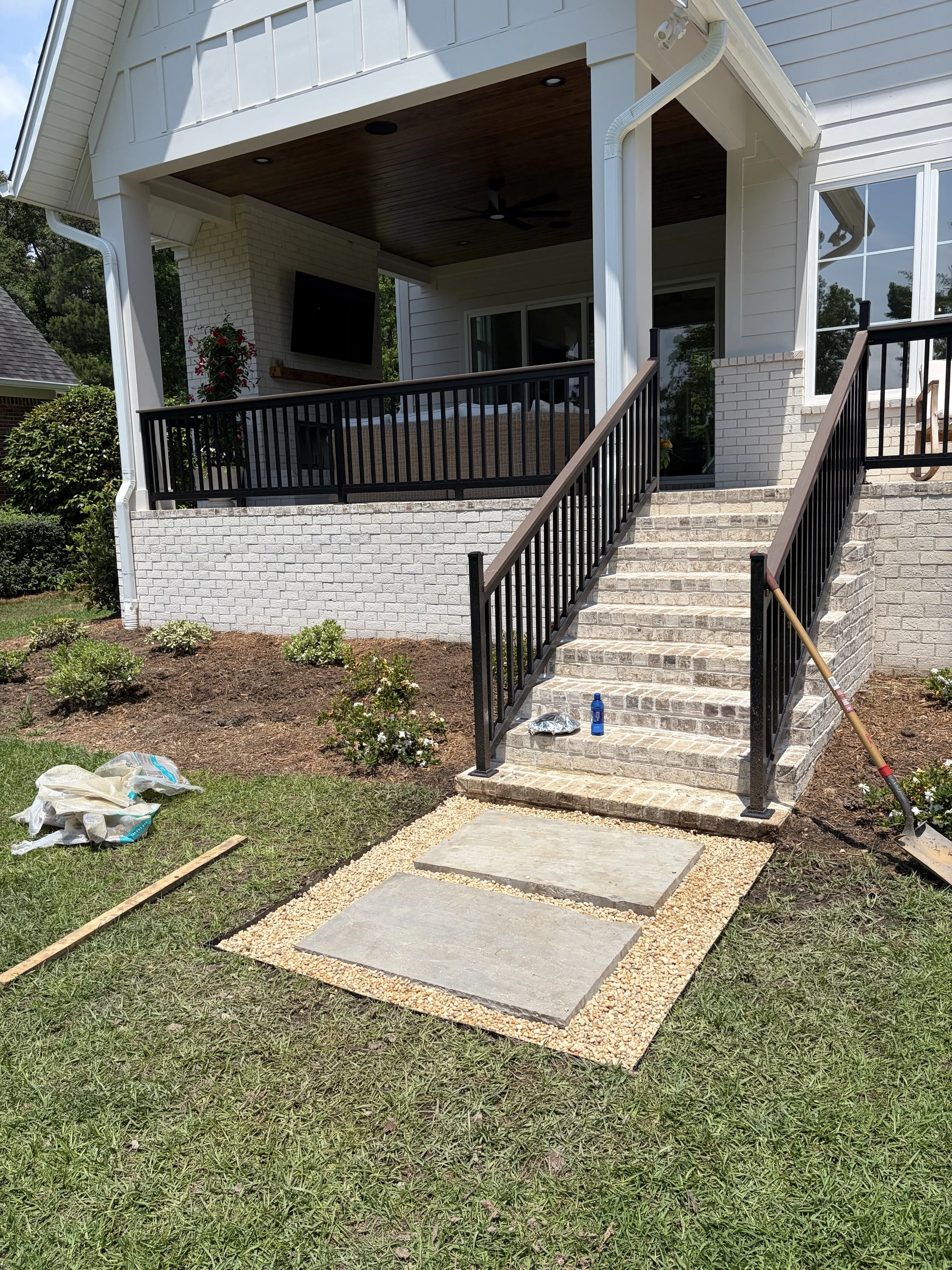 The image shows a house exterior with a small set of brick stairs leading up to a covered porch. The porch has a wood ceiling, a ceiling fan, a mounted TV, and a railing. There are gardening supplies, a shovel, and some potted plants and landscaping around the stairs.