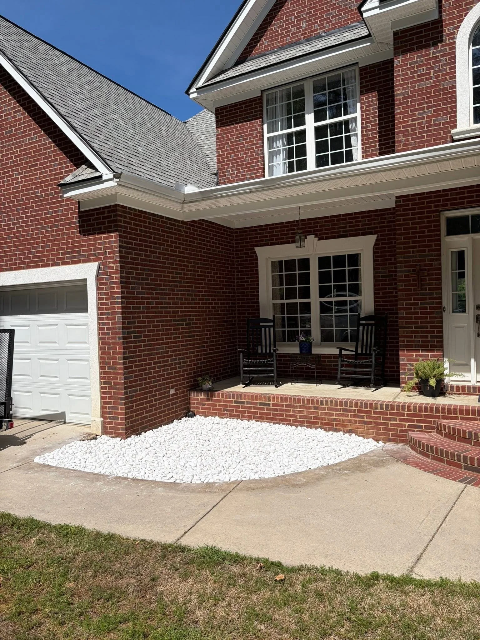 Front view of a brick house with a white garage door, a white porch area with black chairs and potted plants, and a white rock landscape bed, under a clear blue sky.