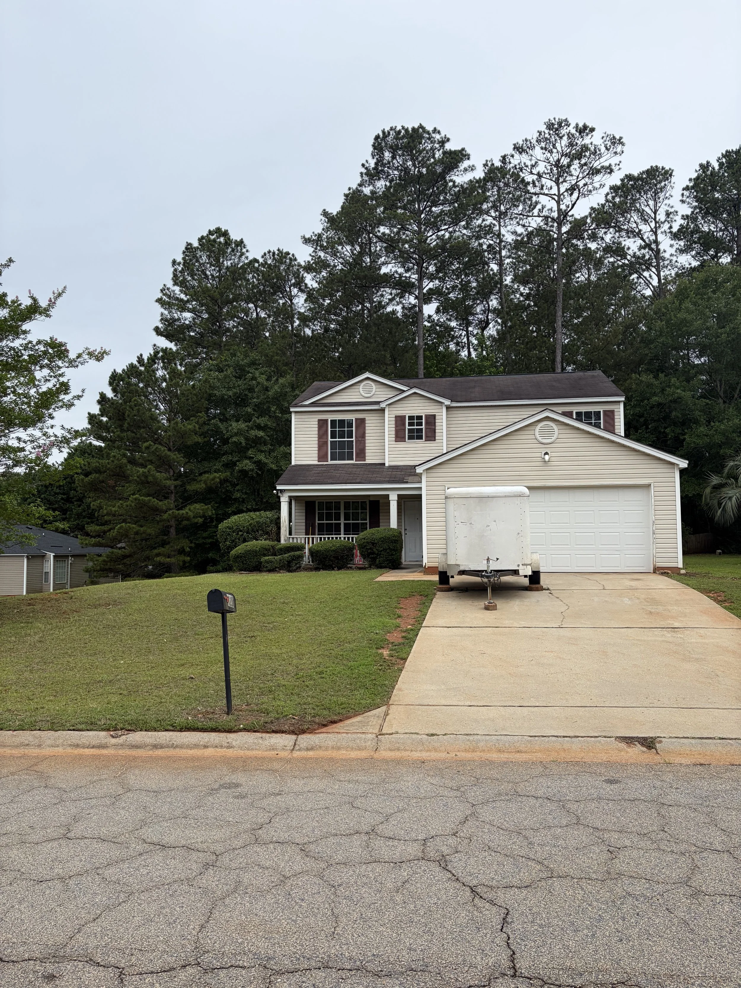 Front yard of a house with a lawn, driveway, and a white trailer parked in front. House has beige siding, brown shutters, and a garage door. Trees and bushes surround the house, and a mailbox is near the street.