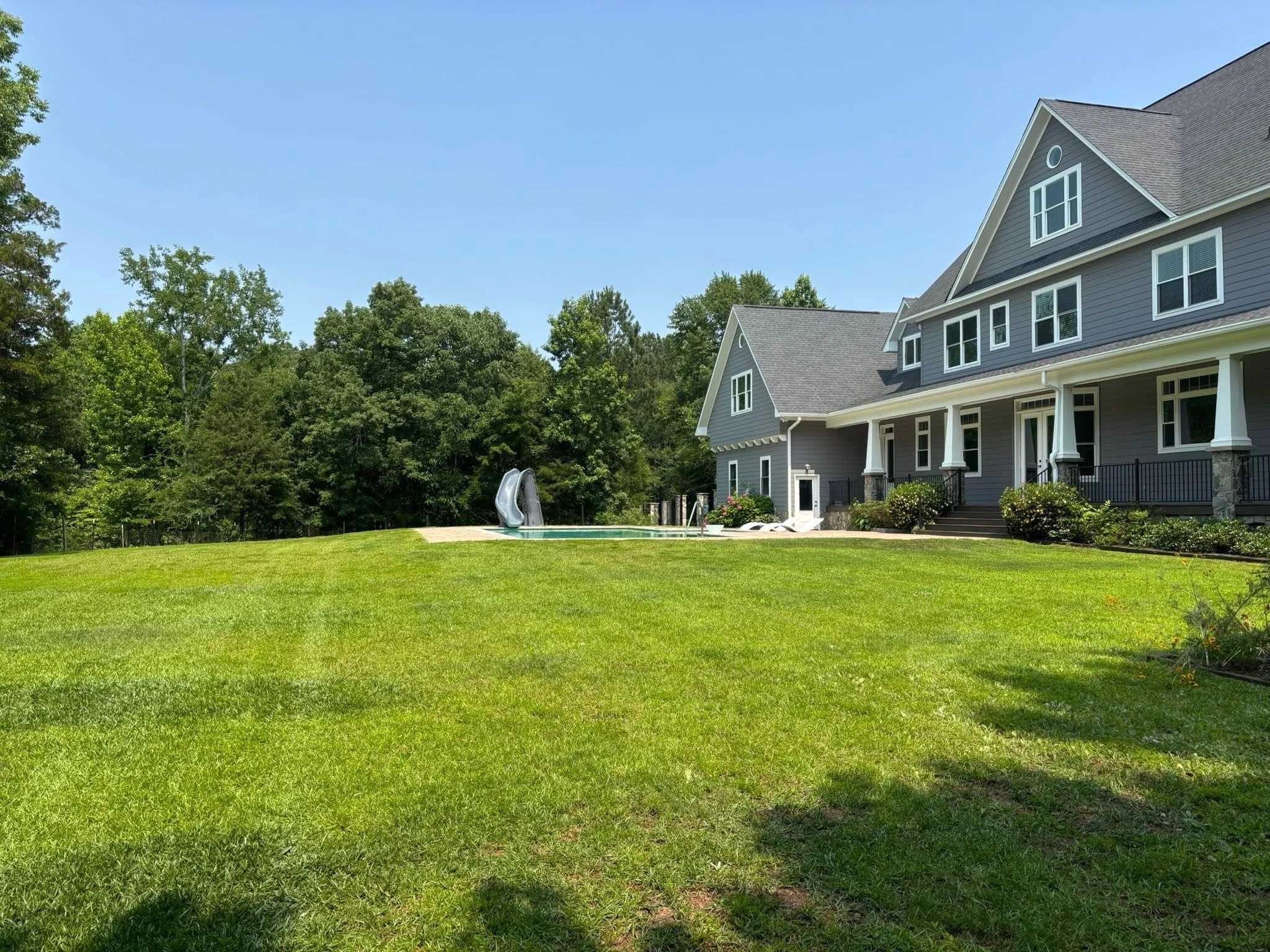 A large gray house with white trim, several windows, and a covered porch, surrounded by green trees and a well-maintained lawn, with a swimming pool and slide in the backyard on a sunny day.