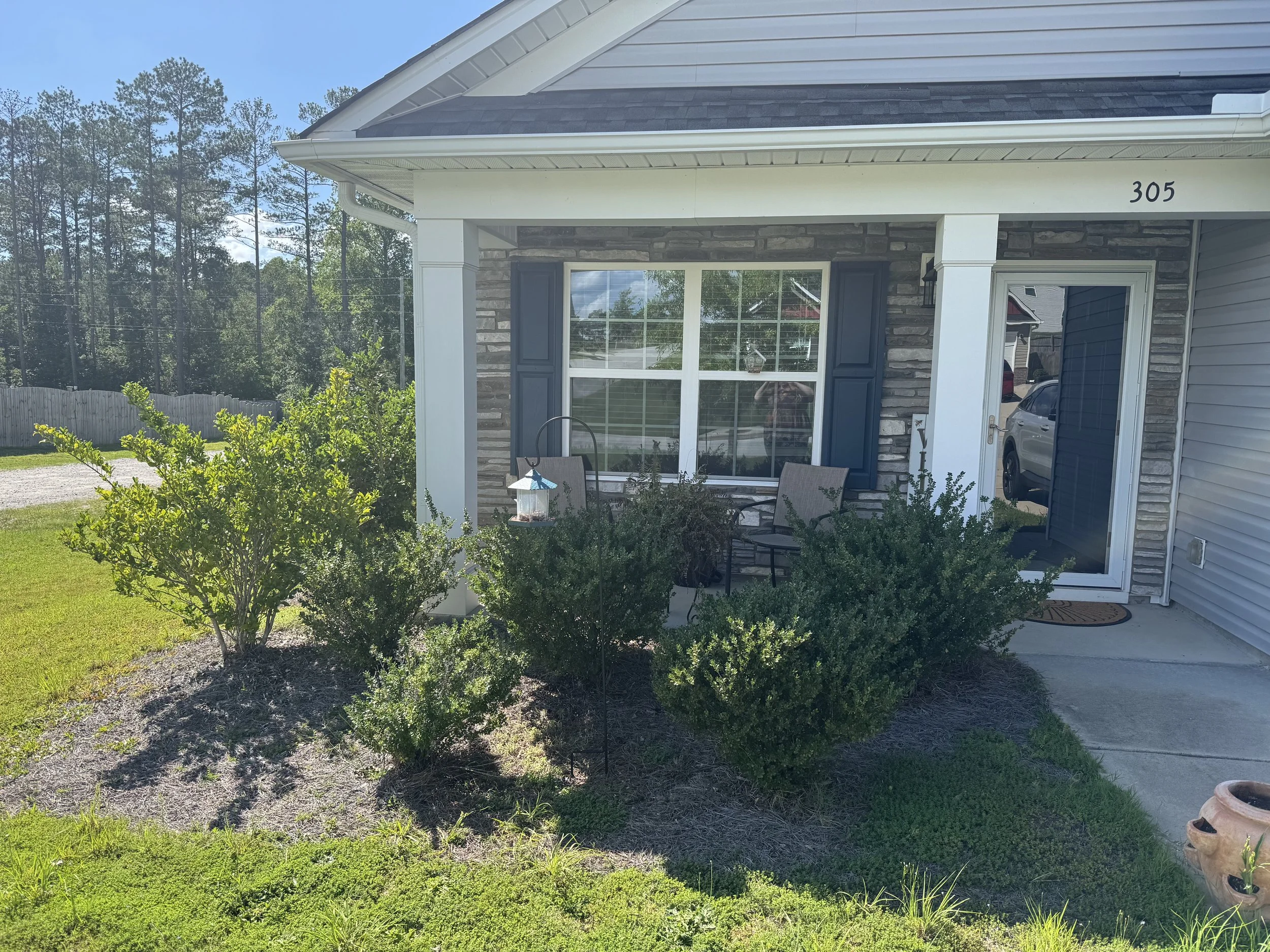 Front porch of a house with large window, blue shutters, white trim, and house number 305. There are chairs and a small table on the porch, and a garden with bushes and a potted plant in front. A white fence and tall trees are visible in the background under a clear blue sky.