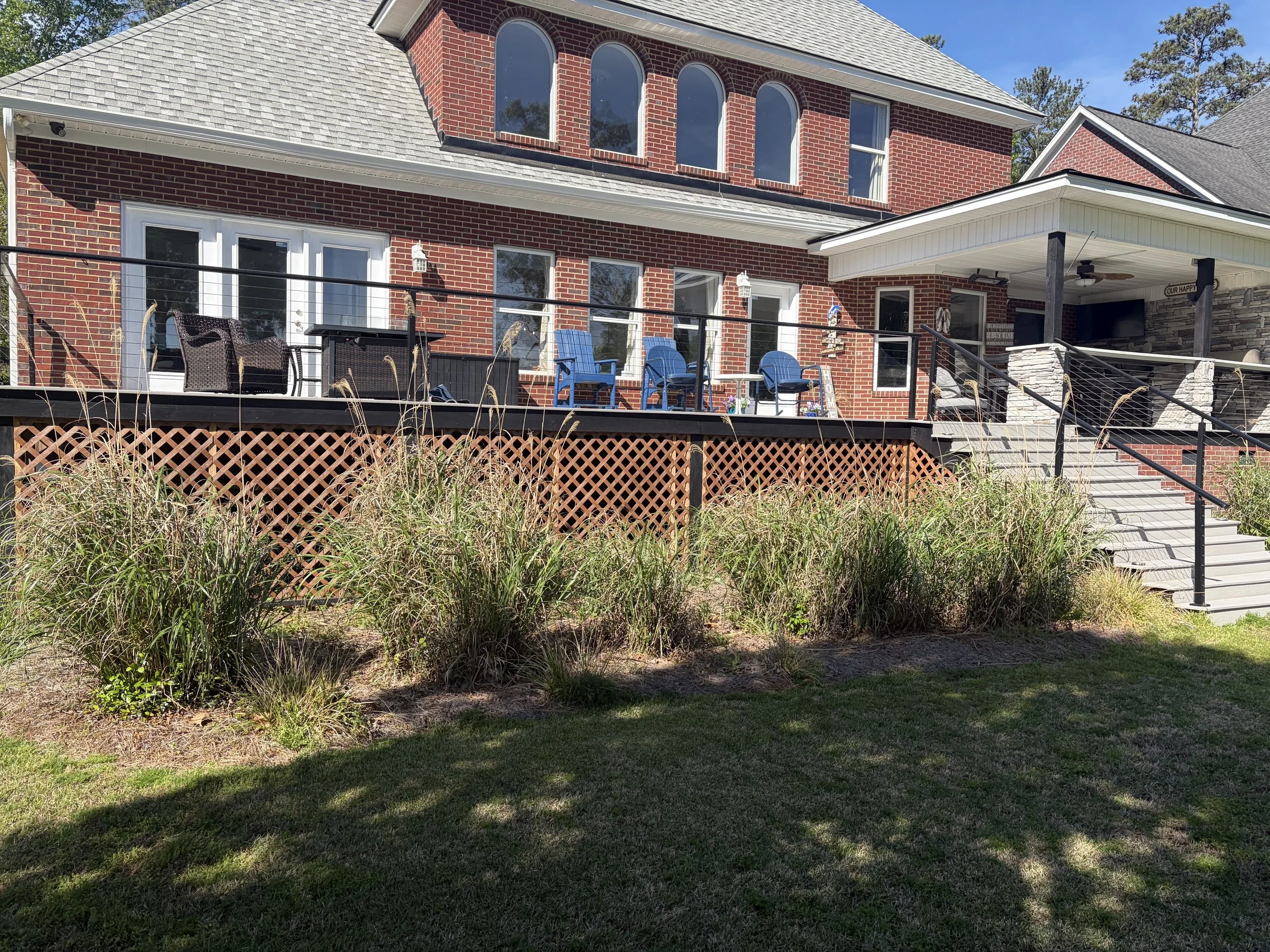 A large brick house with multiple windows and a spacious outdoor deck with chairs and a table. There is a staircase leading up to the deck, and the house has a light-colored roof. The yard has grass and tall ornamental grasses in the foreground, with trees and a clear blue sky in the background.