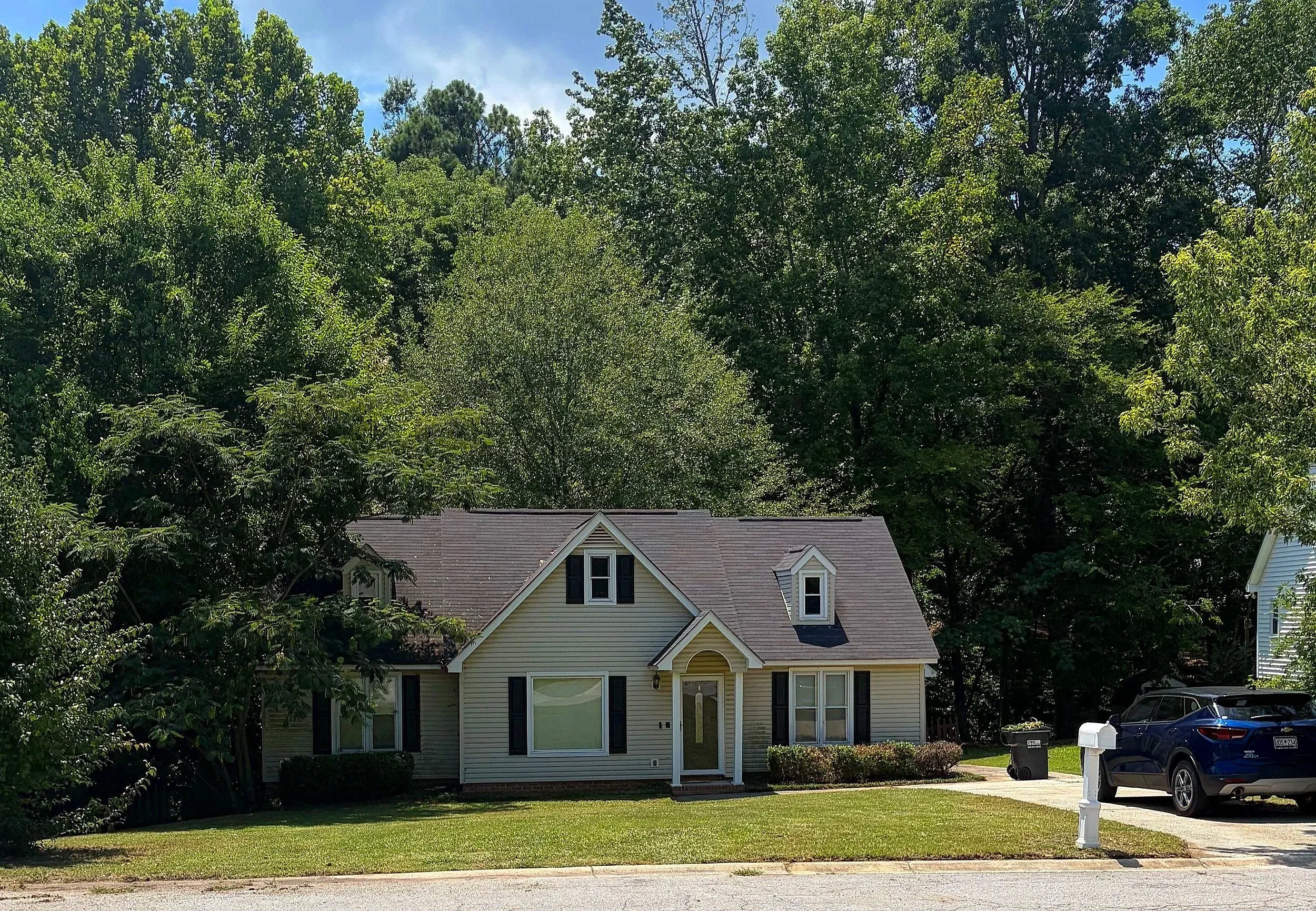 A yellow two-story house with a gray roof and black shutters, surrounded by green trees and a small front lawn. There are parked cars and a mailbox in front of the house on a sunny day.
