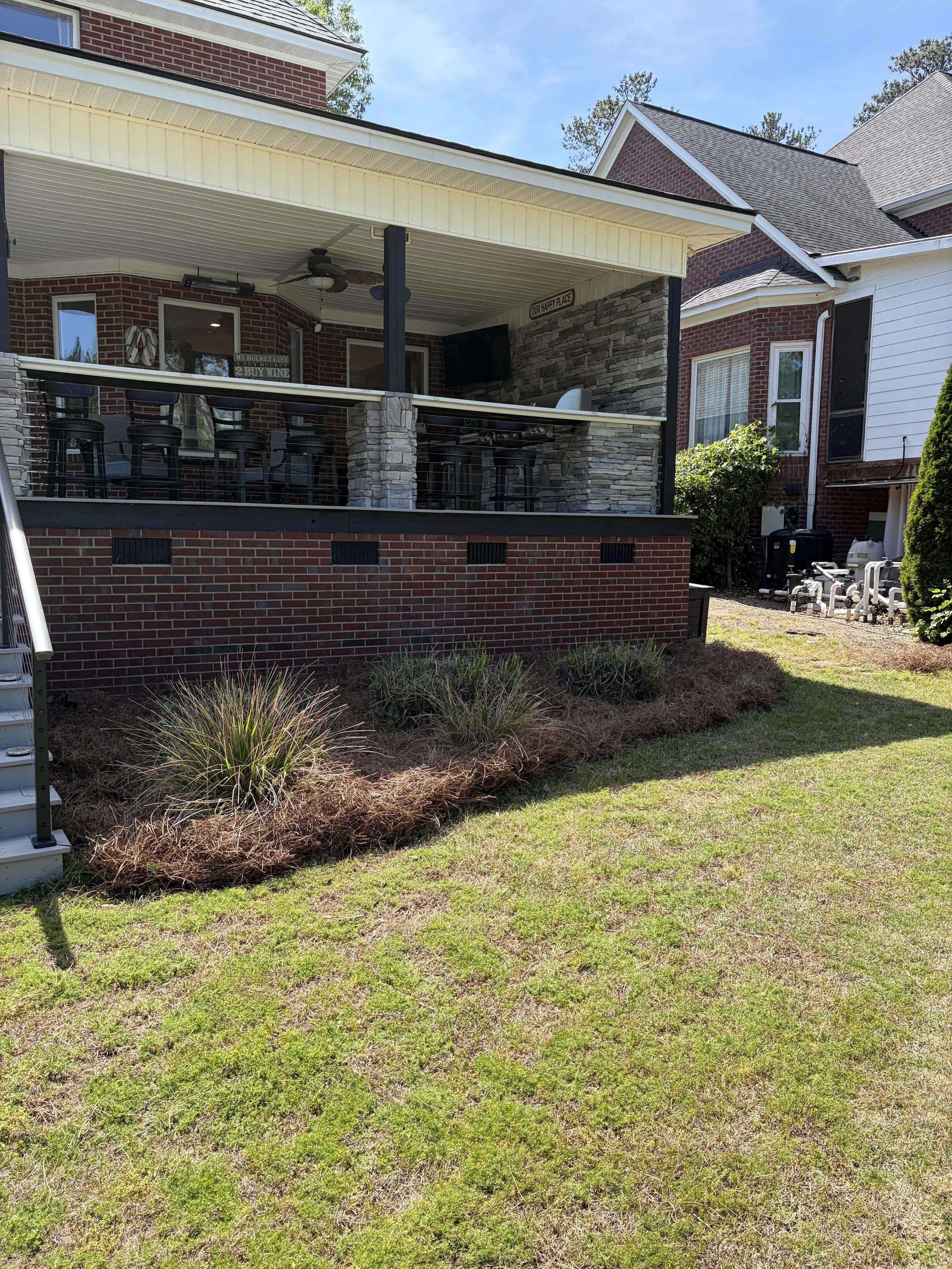Backyard patio with a covered deck, bar stools, a television, a ceiling fan, and some surrounding plants and grass.