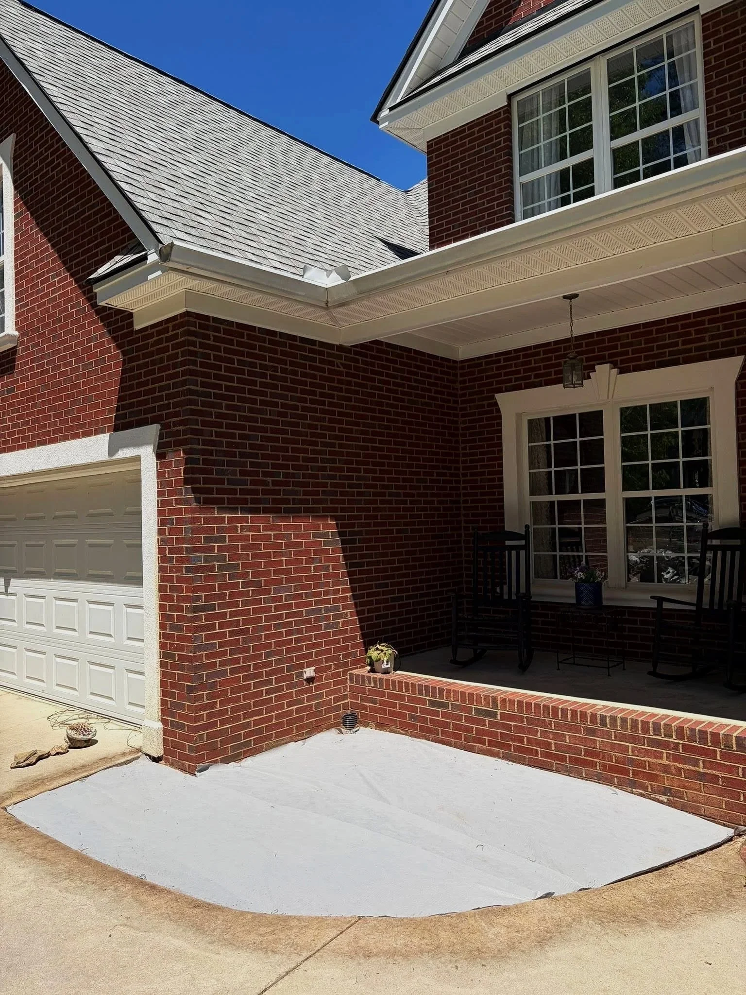 Close-up of a brick house exterior with a white garage door, sliding glass door with black chairs and potted plants on the porch, and a freshly painted concrete patio surface.
