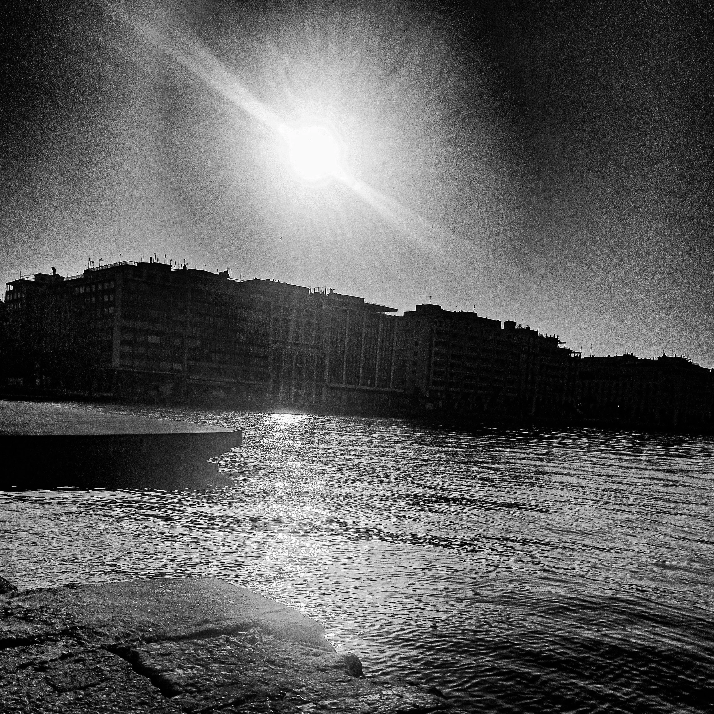 A black and white photo of buildings along the shoreline, with the sun reflecting on water and a bird flying in the sky.