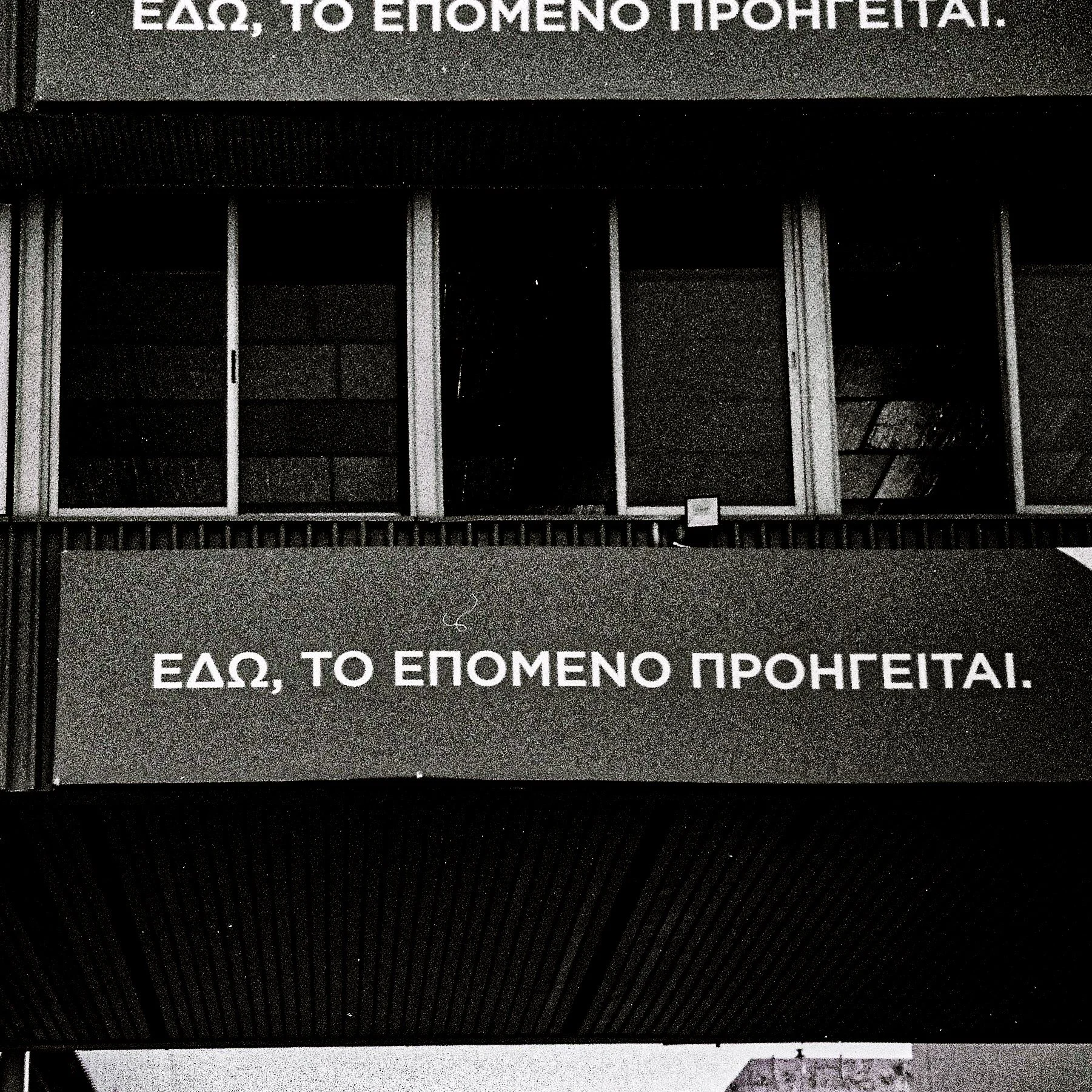 A close-up of a black and gray sign with Greek letters and a line of text that reads, 'ΕΔΩ, ΤΟ ΕΠΟΜΕΝΟ ΠΡΟΗΓΕΙΤΑΙ!'