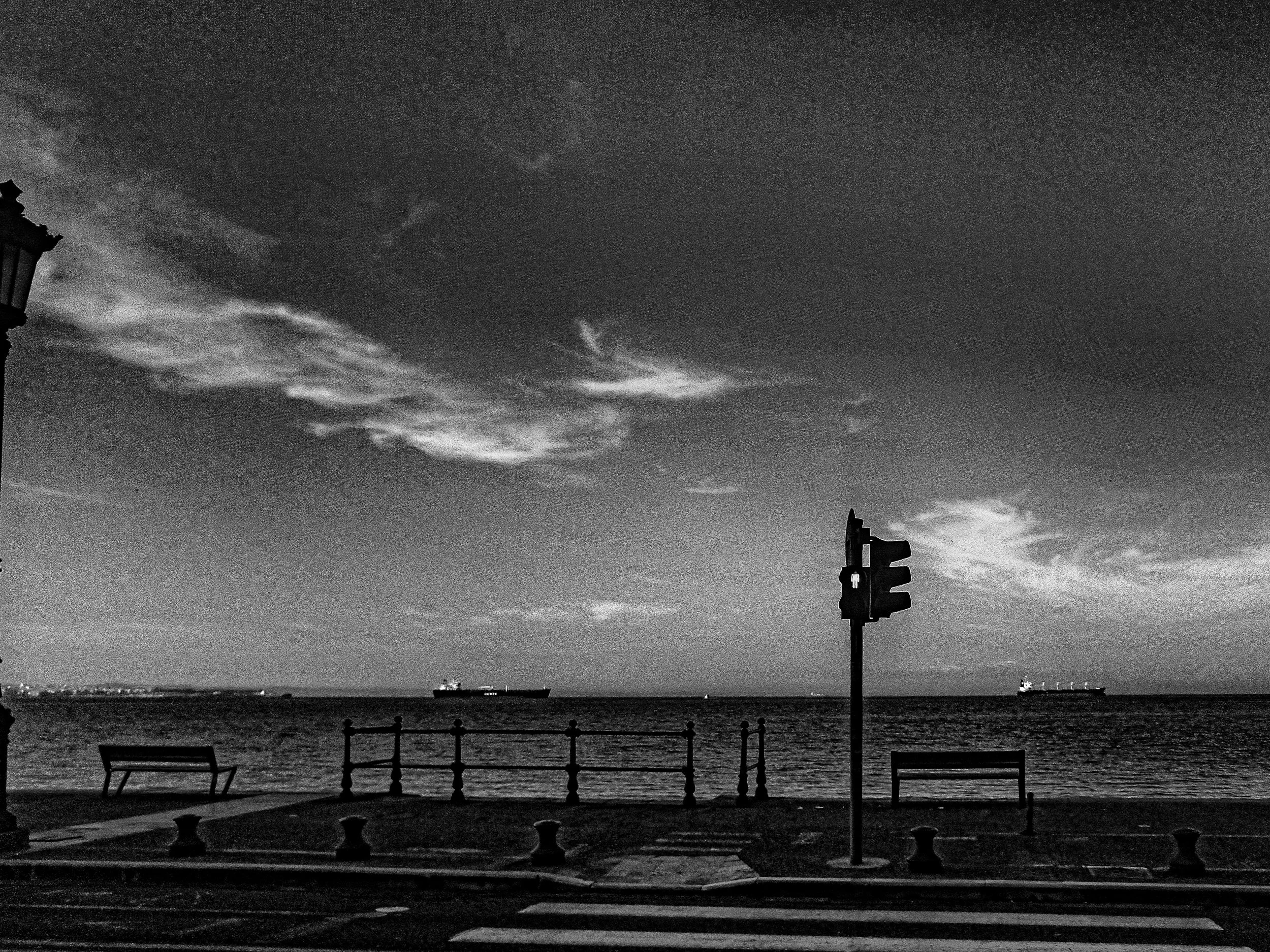 Black and white photograph of a nighttime scene at the waterfront with benches, a lamp post, ships on the horizon, and a mostly cloudy sky.