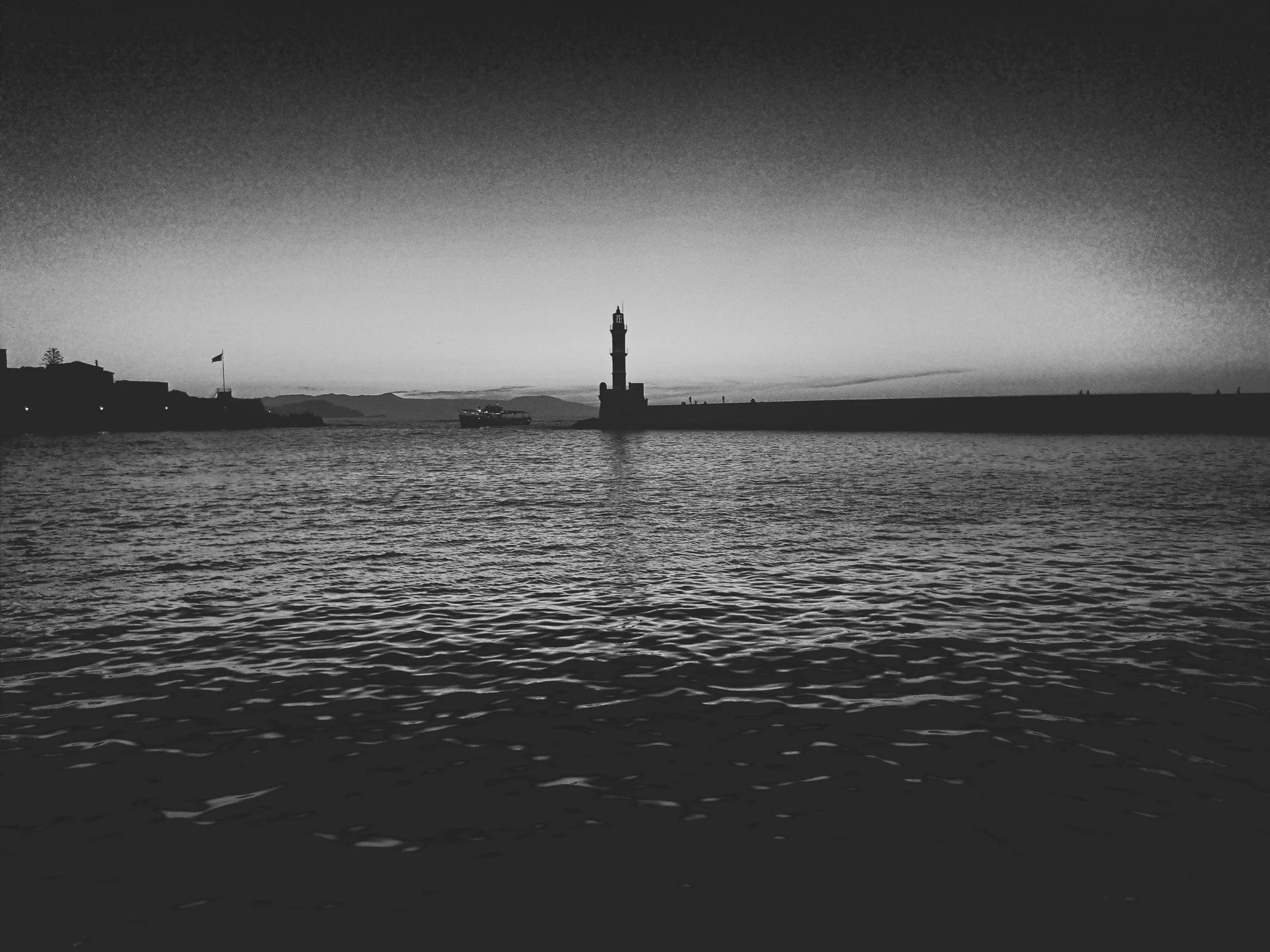 A black and white photo of a lighthouse on a breakwater, with a boat nearby and a distant shoreline with hills.