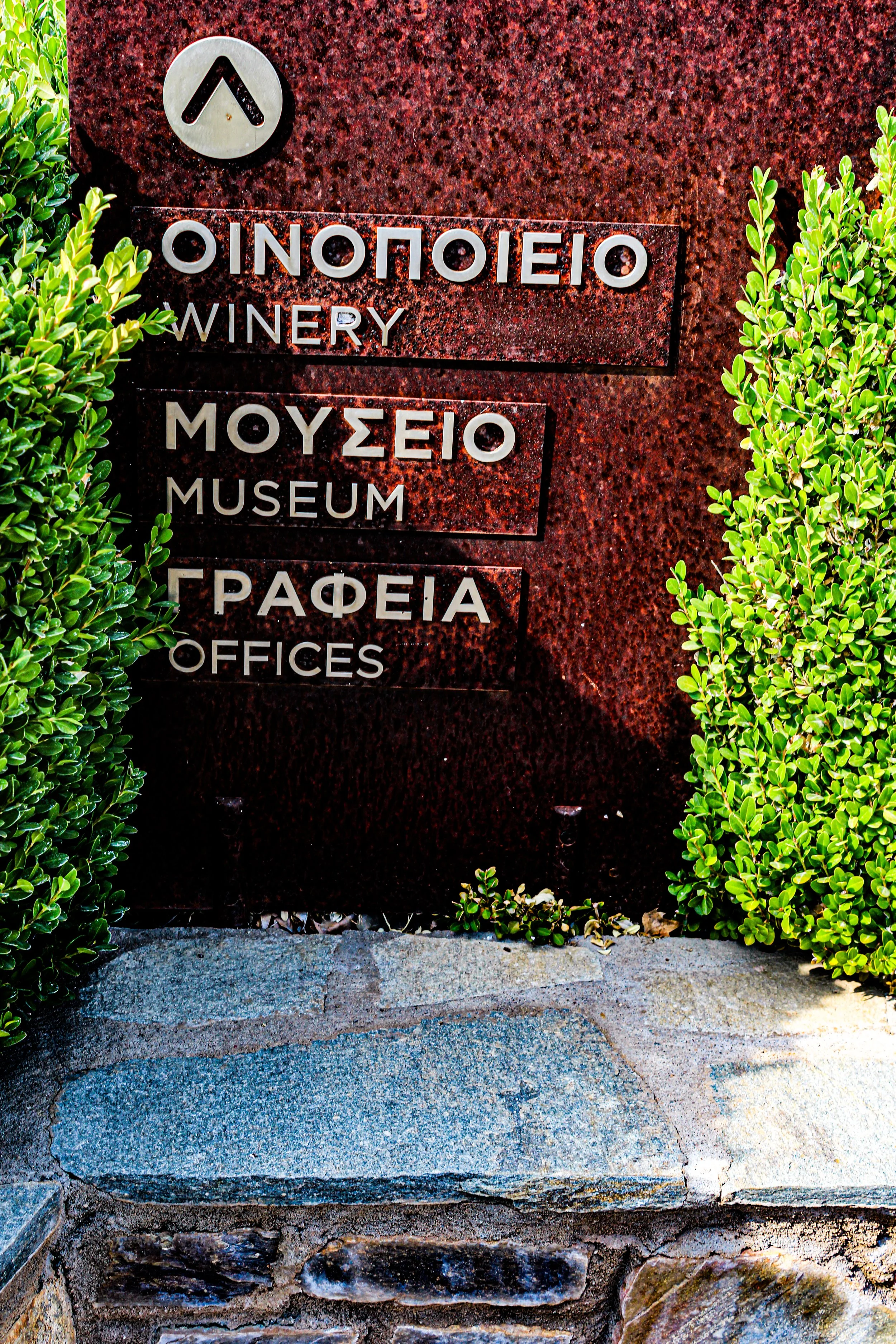 Sign with Greek and English text indicating a winery, museum, and offices, surrounded by green bushes and positioned on a red marble wall, with stone steps in front.