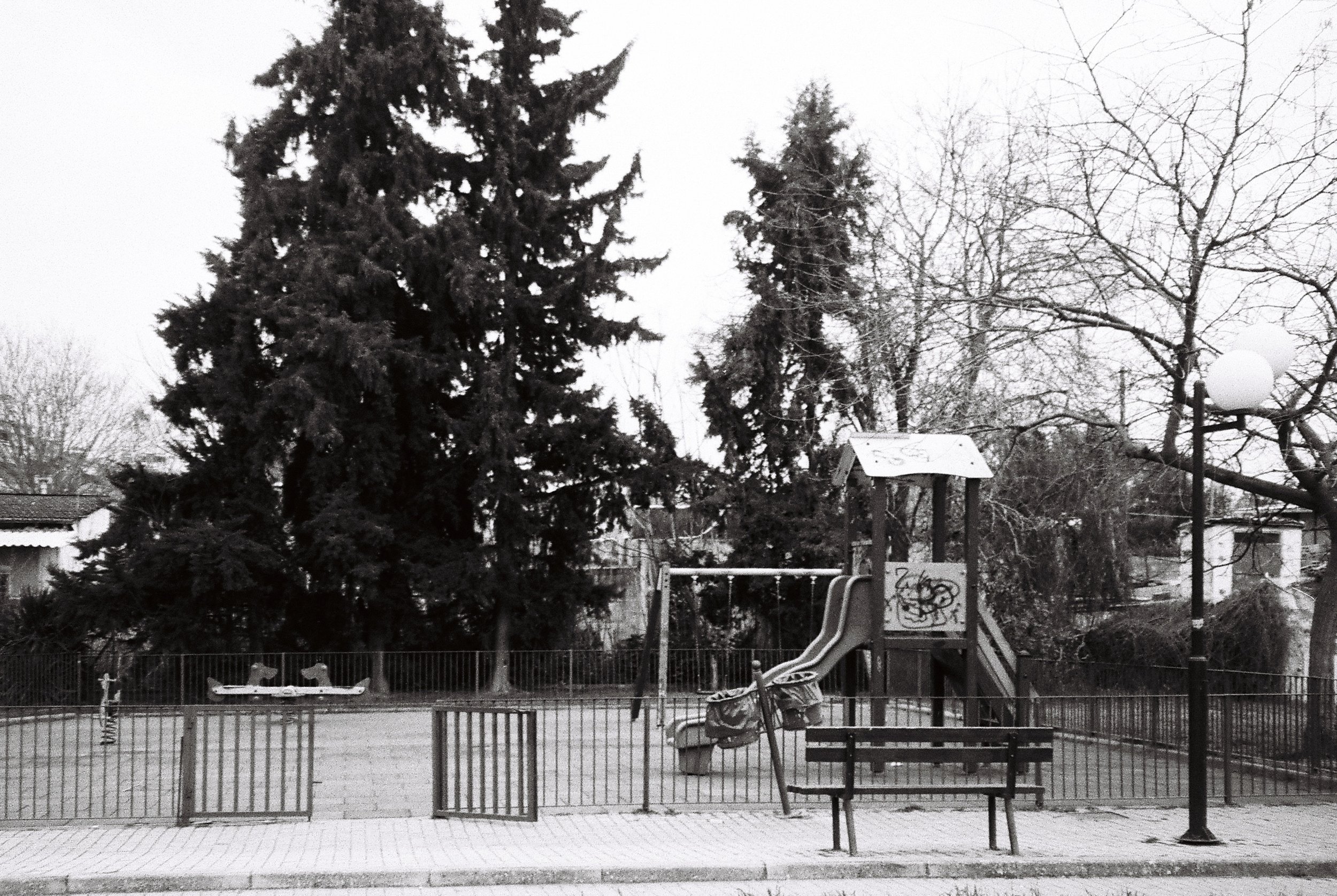 A playground enclosed by a metal fence, featuring a play structure with a slide, a small climbing wall, and swings, set against a background of leafless trees and tall evergreen trees, with overcast sky.