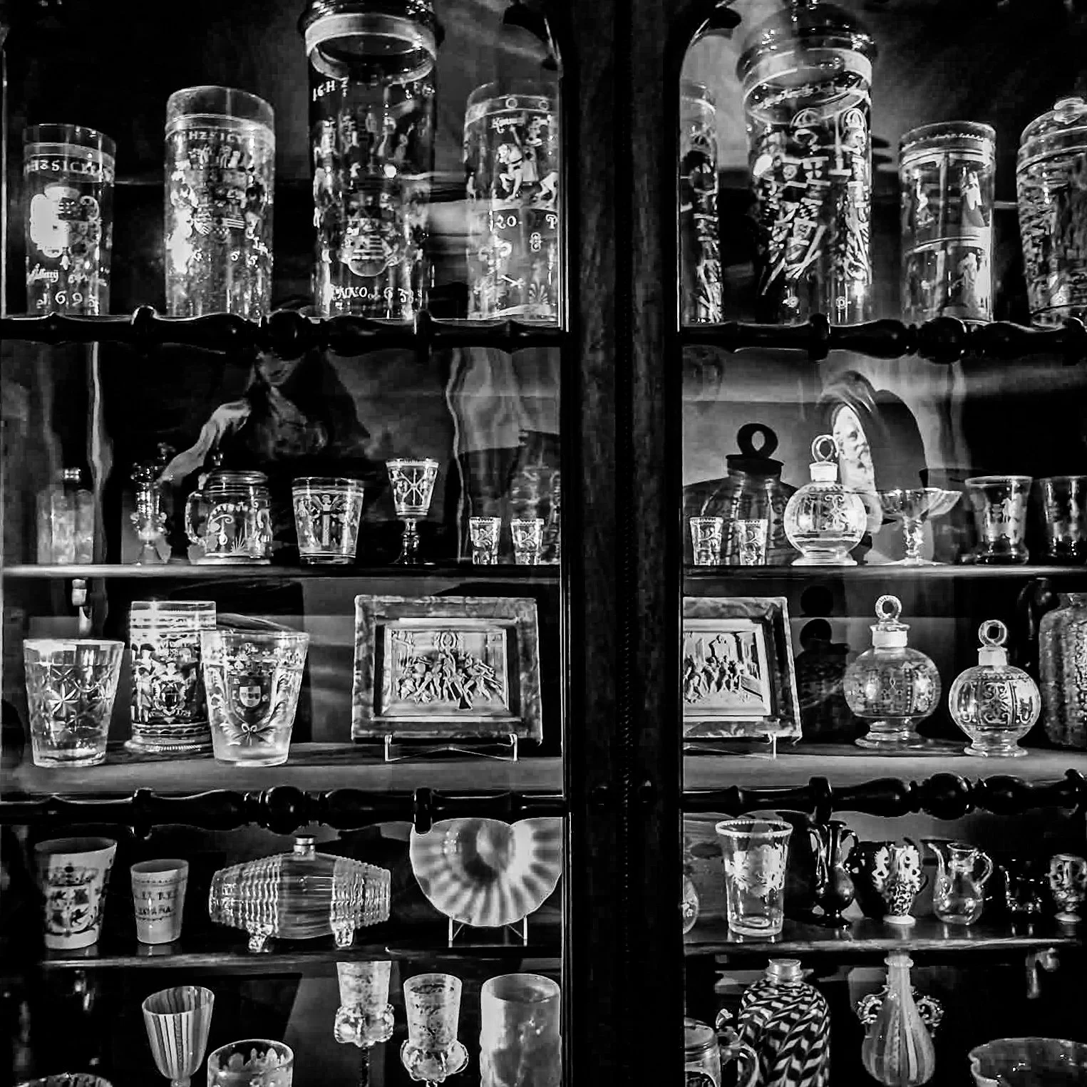 Display of various glasses, vases, and decorative items arranged on a wooden cabinet with glass doors, including carved and patterned glassware and framed artwork, in black and white.