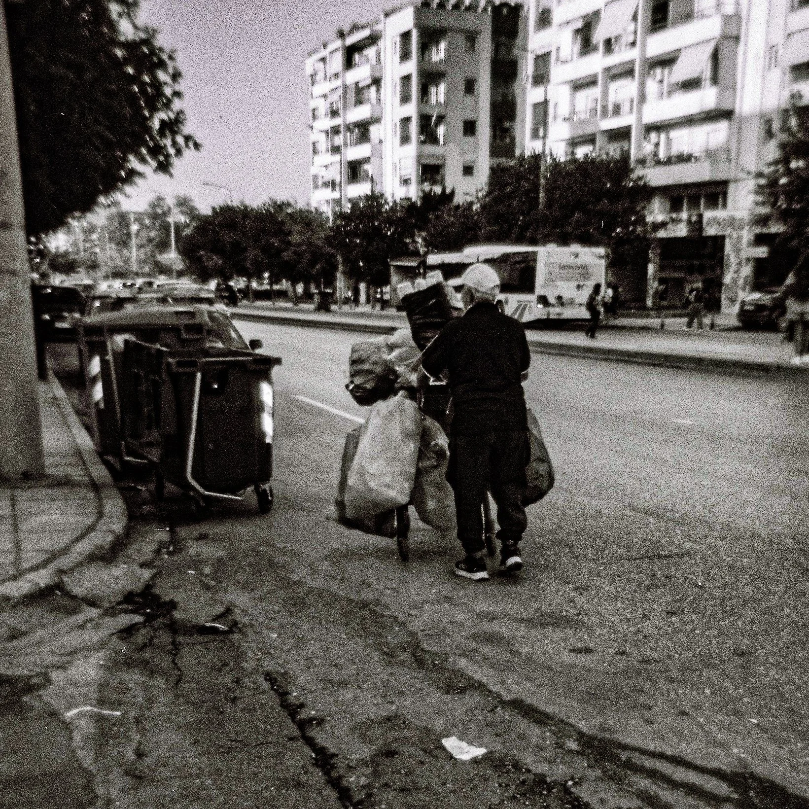 A black and white photo of a city street with high-rise buildings, some trees, and pedestrians in the background. Two people are walking with luggage, and a delivery truck is visible on the street.