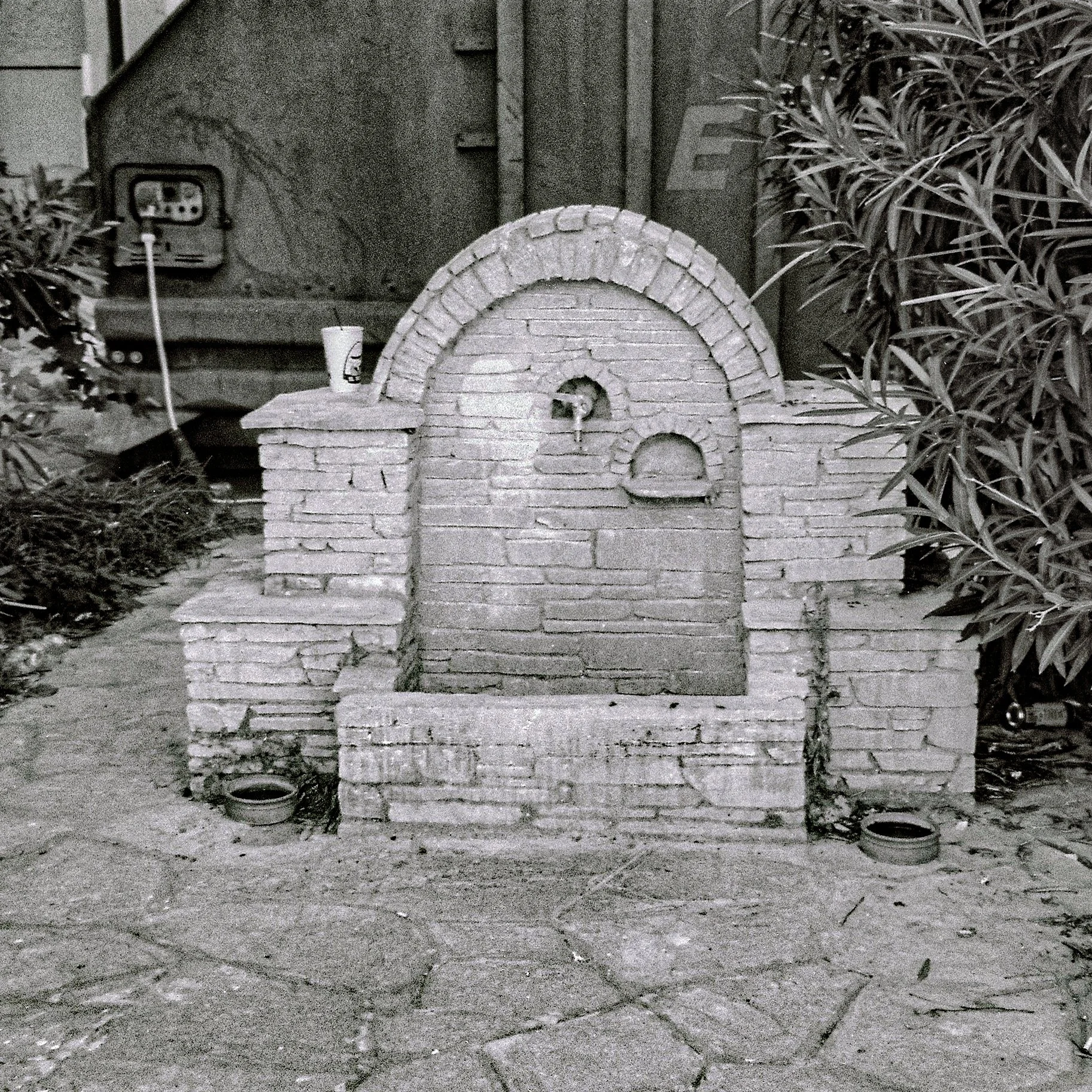 A vintage-style brick pizza oven with a chimney, set outdoors on a paved area with plants on either side and an electrical box mounted on a wall in the background.