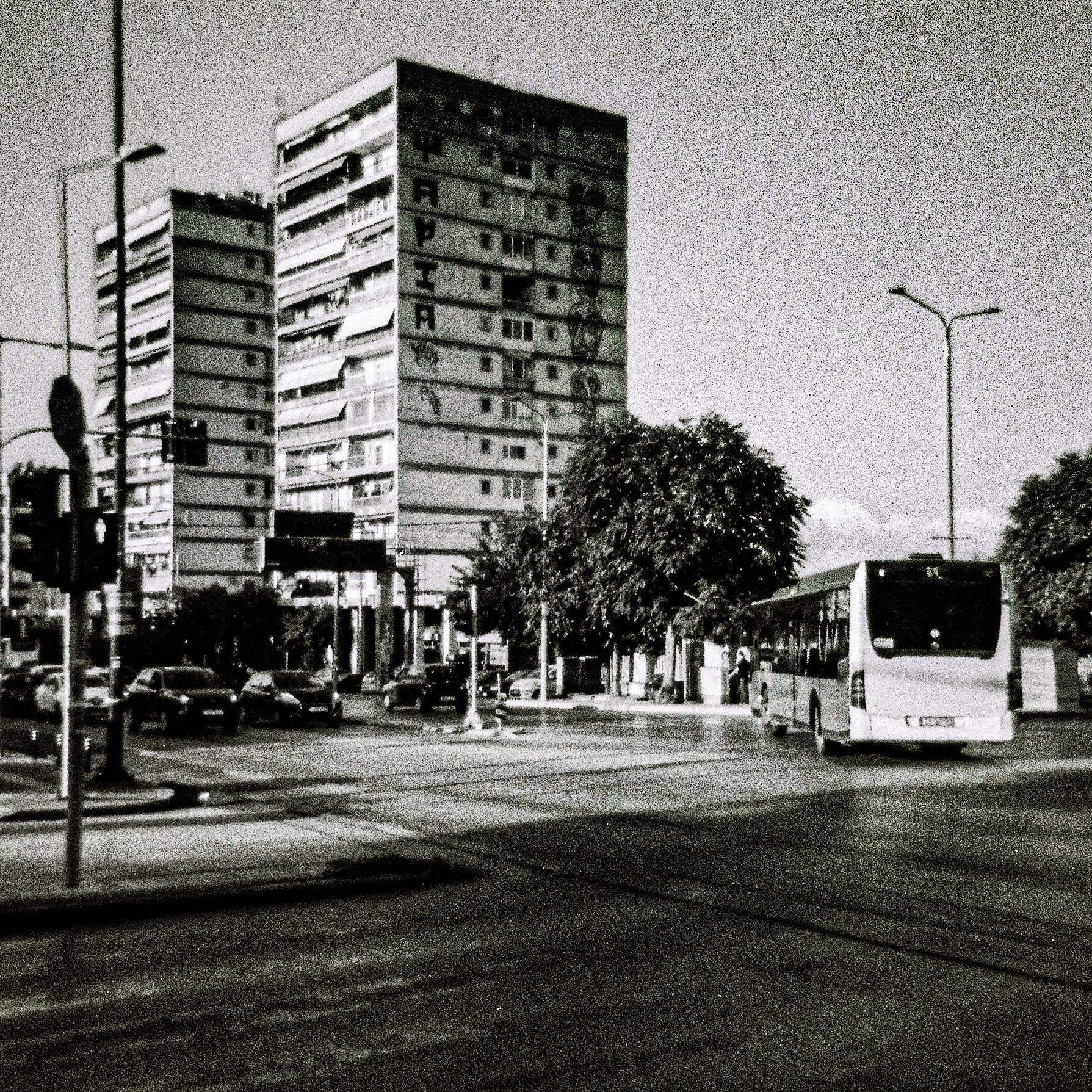 Black and white photo of a city street scene during the night with high-rise buildings, parked cars, a bus, streetlights, and pedestrians.