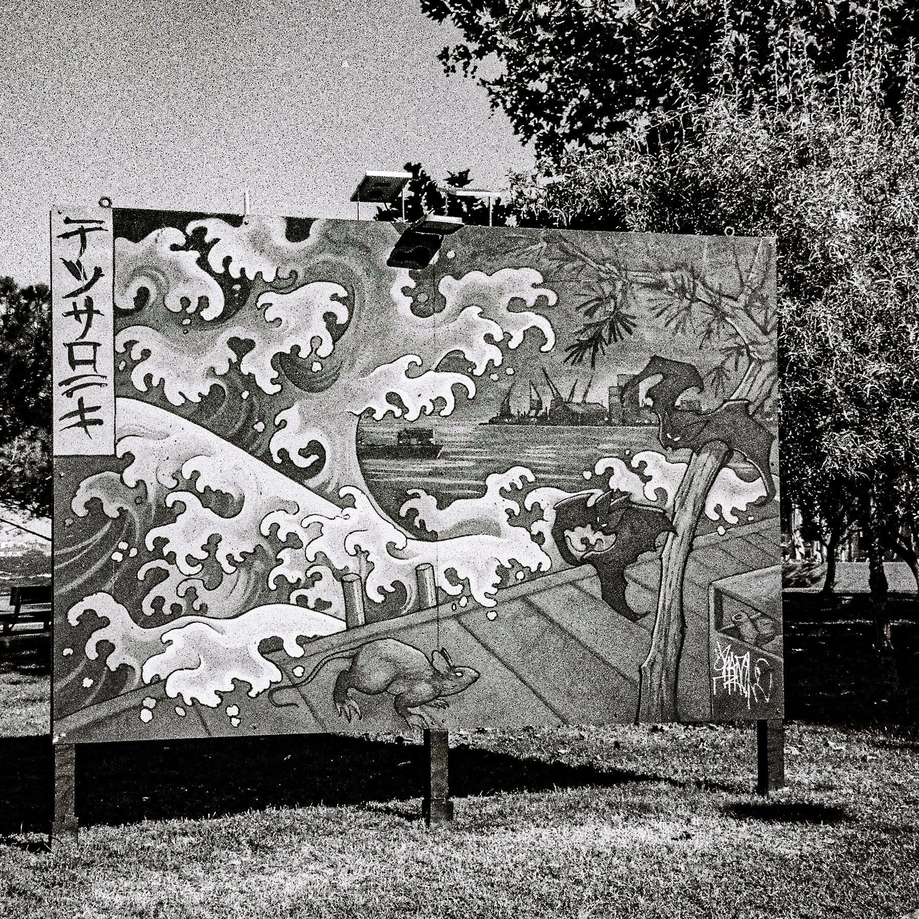 Black and white mural of a dragon surrounded by clouds and water, with Japanese characters on the left side, outdoors with trees and gravel at the base.