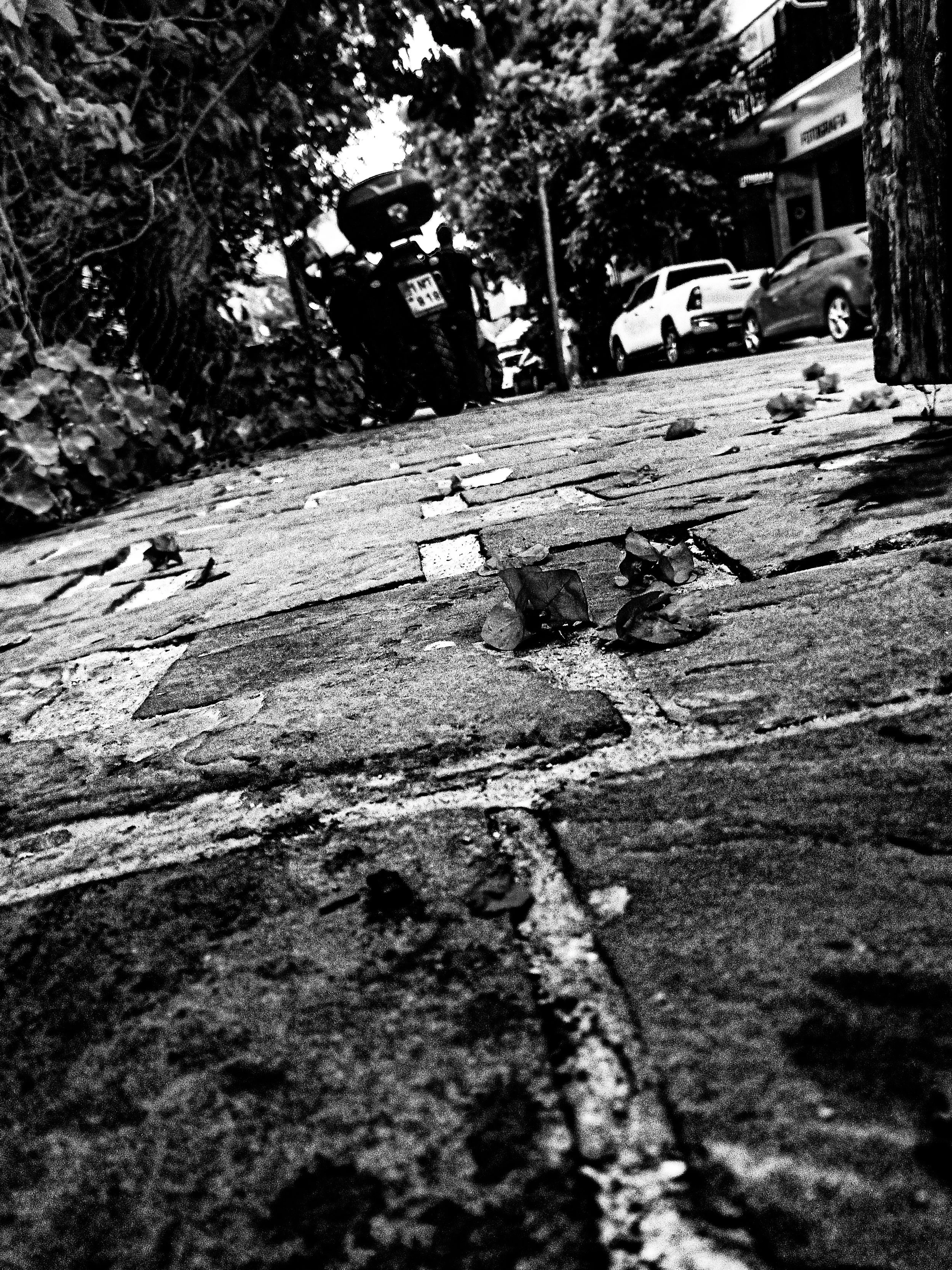A low-angle black and white photograph of a city sidewalk with fallen leaves. In the background, parked cars and trees are visible, along with a person standing near a motorcycle.