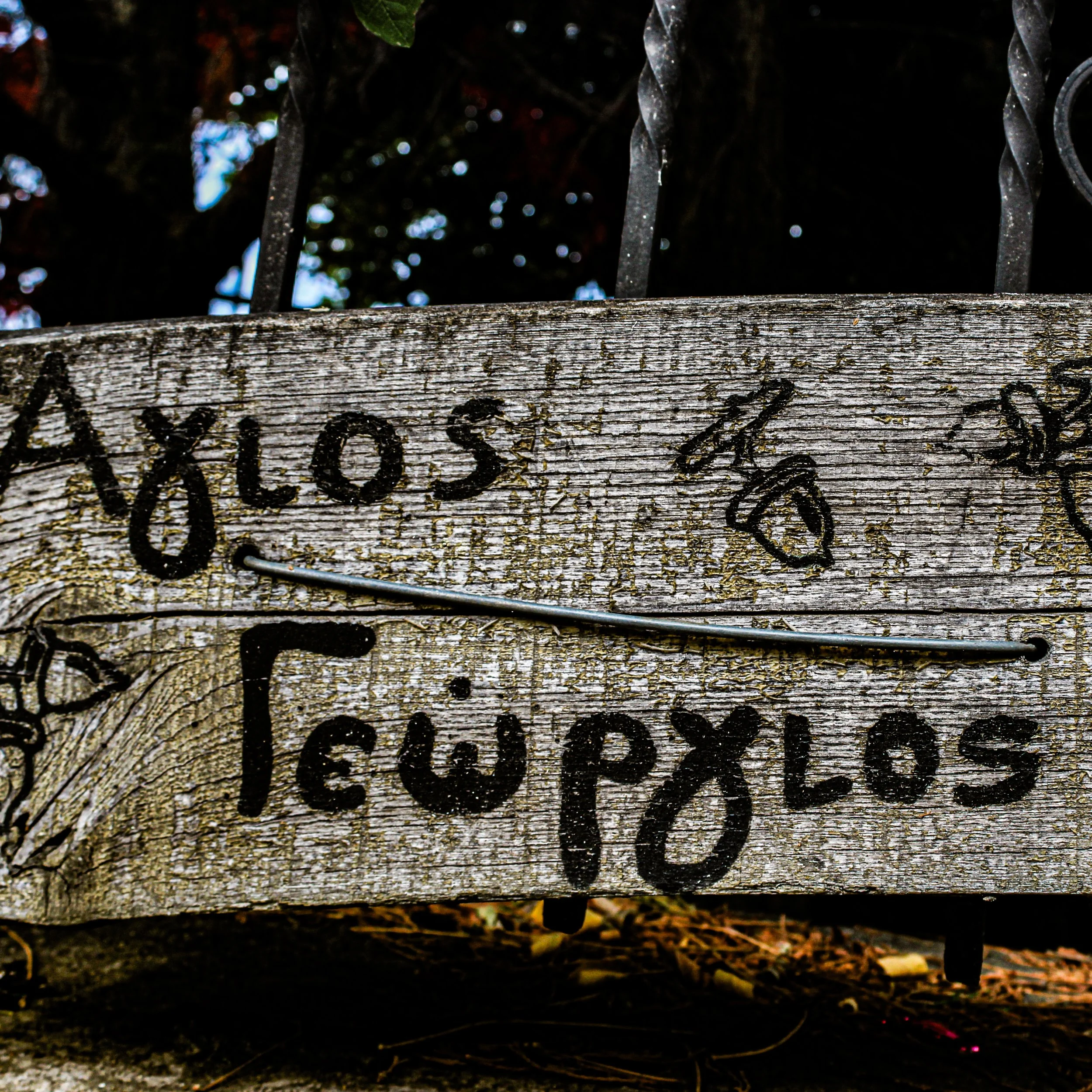 Close-up of a weathered wooden sign with black handwritten text, partially visible, beneath a black wrought iron fence against a dark background with some blurred lights.
