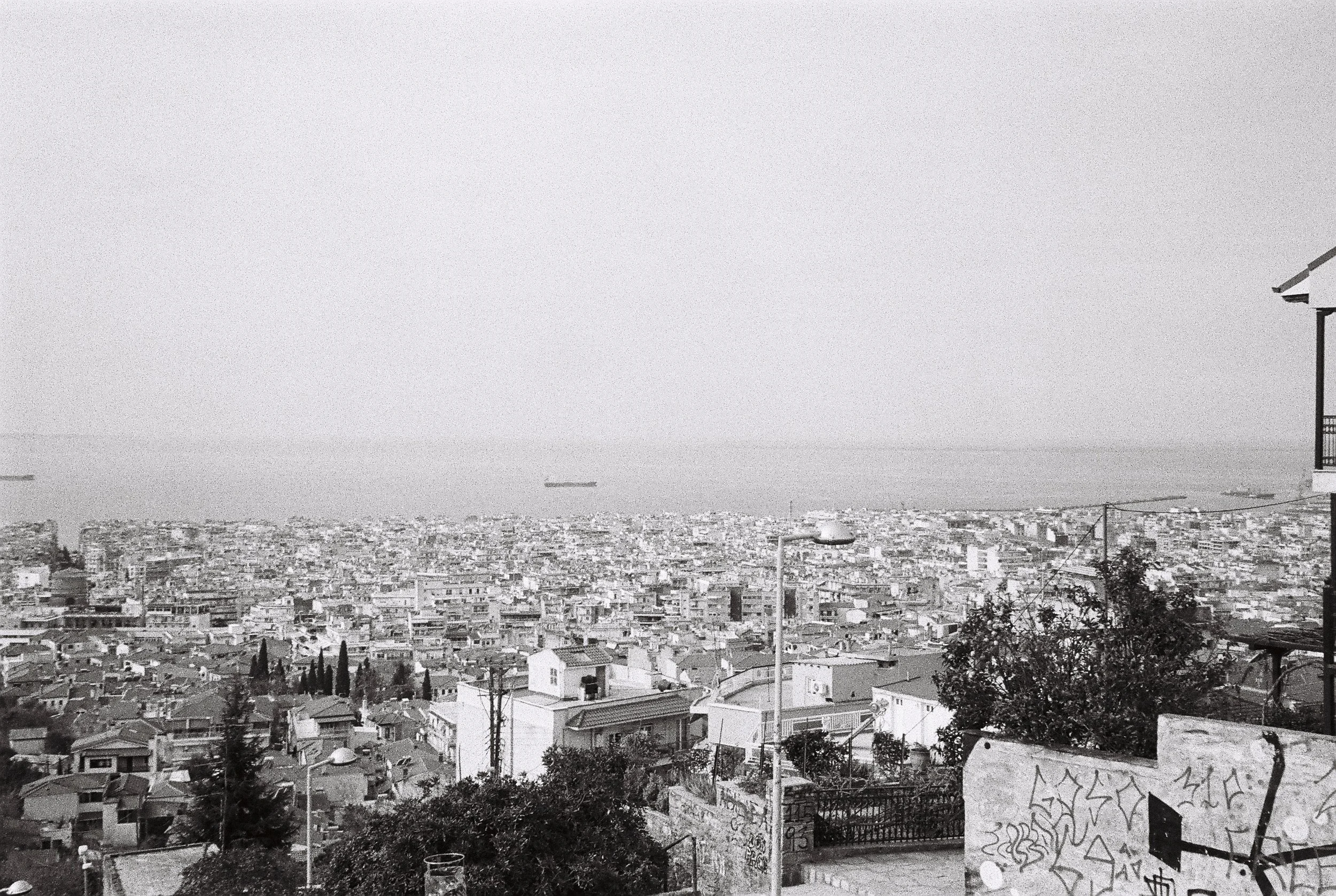 A black and white photo of a cityscape with numerous buildings extending to the horizon, a large body of water in the distance with cargo ships, and a street lamp and graffiti-covered wall in the foreground.